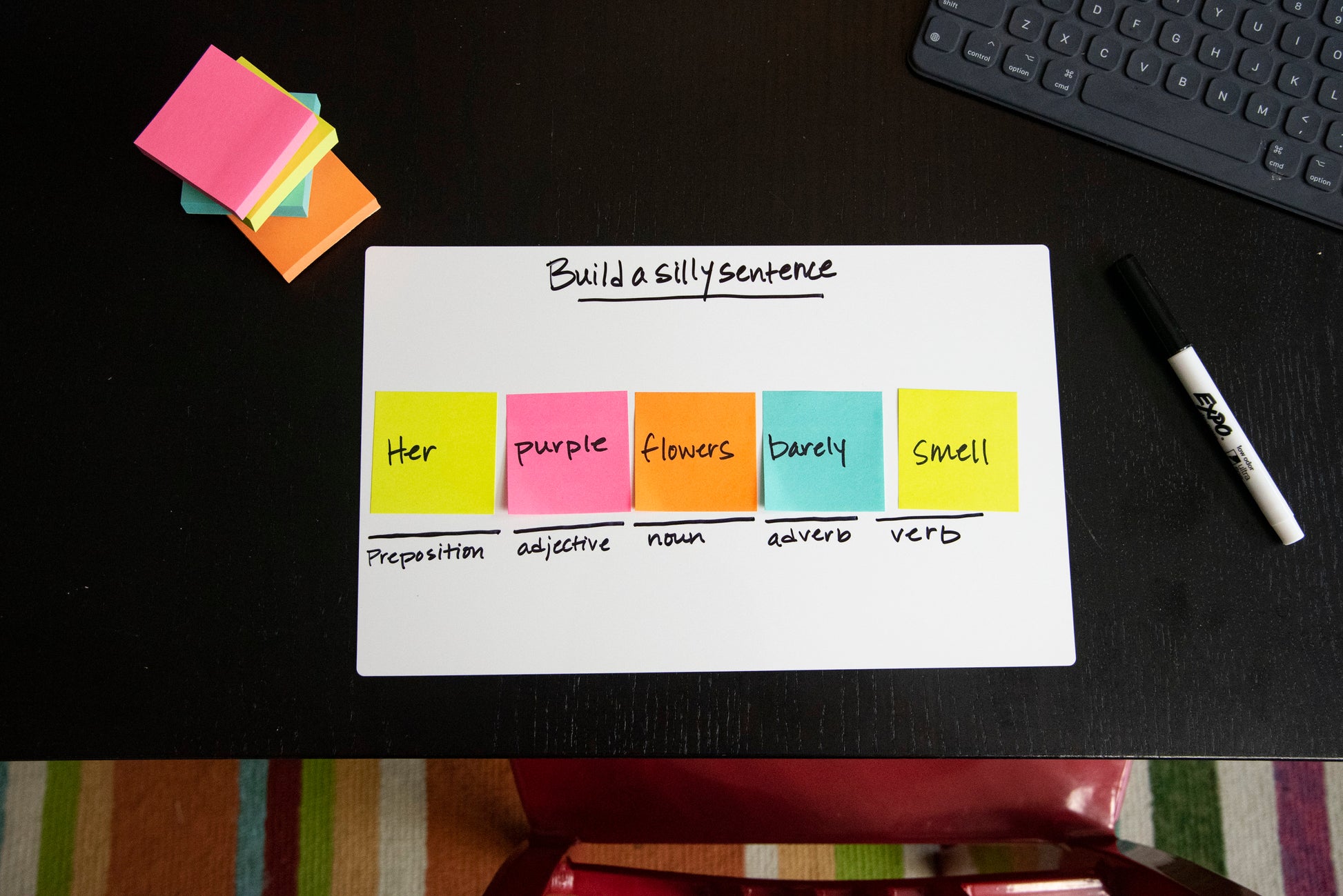 Ein Post-it® Notes Würfel der 3M Deutschland GmbH (76x76mm, 450 Blatt, 100% PEFC) zeigt eine Satzbauaufgabe auf einem Whiteboard mit Grammatiketiketiketten; zusätzliche Haftnotizen, ein Marker, eine Tastatur und ein gestreifter Teppich liegen in der Nähe.