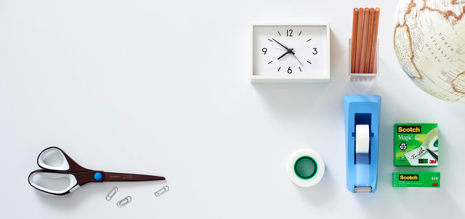 Eine Scotch™ Titanschere (20 cm) von 3M Deutschland GmbH, drei Büroklammern, eine weiße Uhr, ein Bleistiftbehälter, ein Globus, ein Klebebandabroller und eine Schachtel Scotch-Klebeband sind ordentlich auf einer weißen Fläche angeordnet.