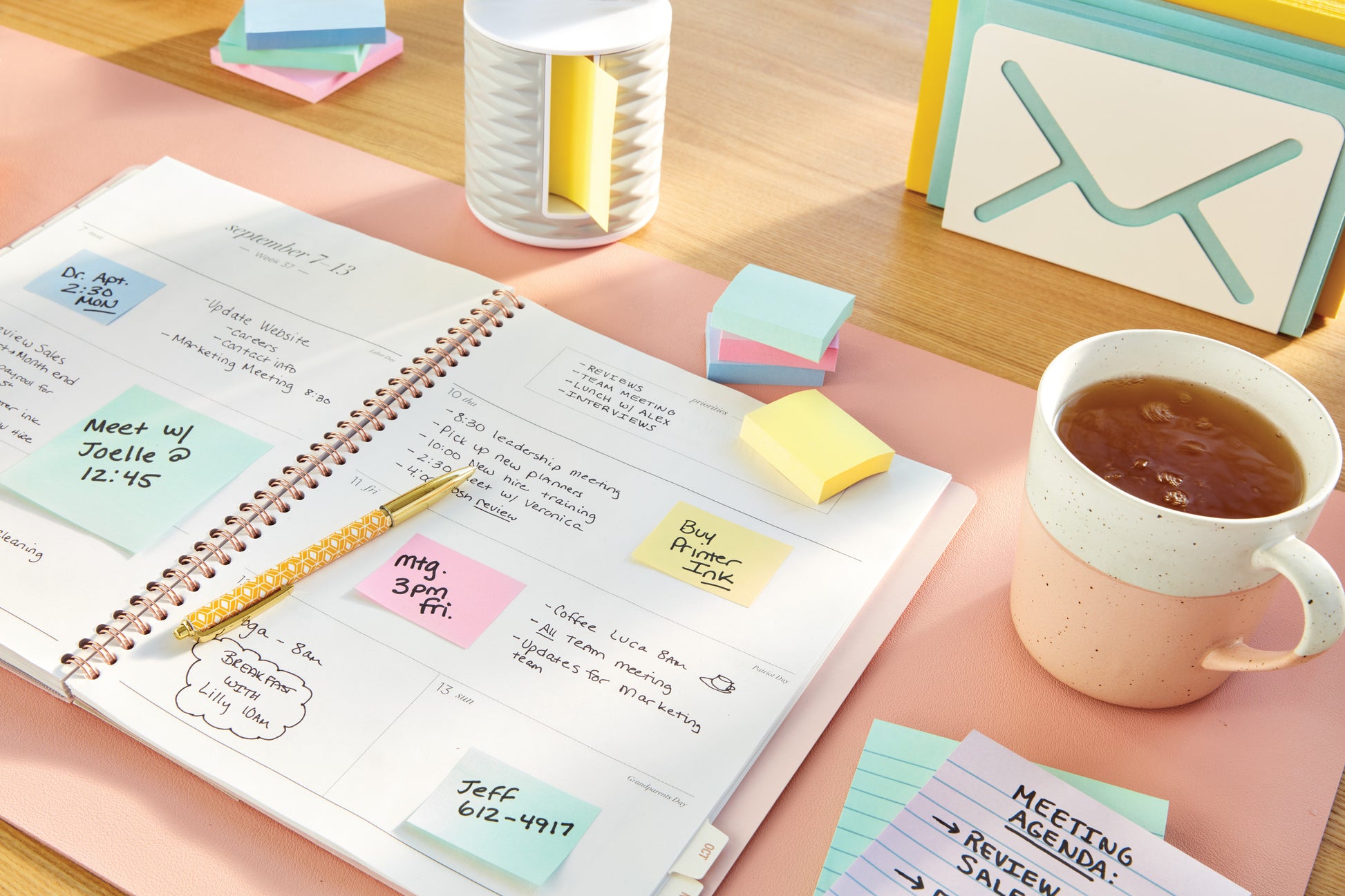 Ein offener Planer mit handschriftlichen Notizen liegt auf einem Schreibtisch, umgeben von einer Tasse Tee, einem Stift, einem dekorativen Organizer, Karteikarten mit der Aufschrift "Meeting Agenda" und Post-it® Notes (Gelb, 38 x 51 mm) der 3M Deutschland GmbH.