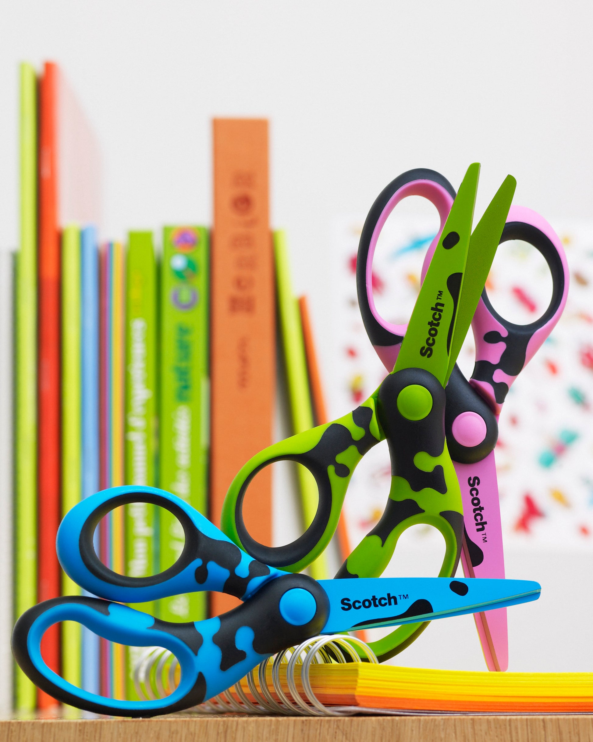 Zwei Scotch™ DECO Kinderscheren (grün und blau), je 13 cm von 3M Deutschland GmbH, sind auf buntem Papier mit aufrecht stehenden Büchern im Hintergrund platziert.