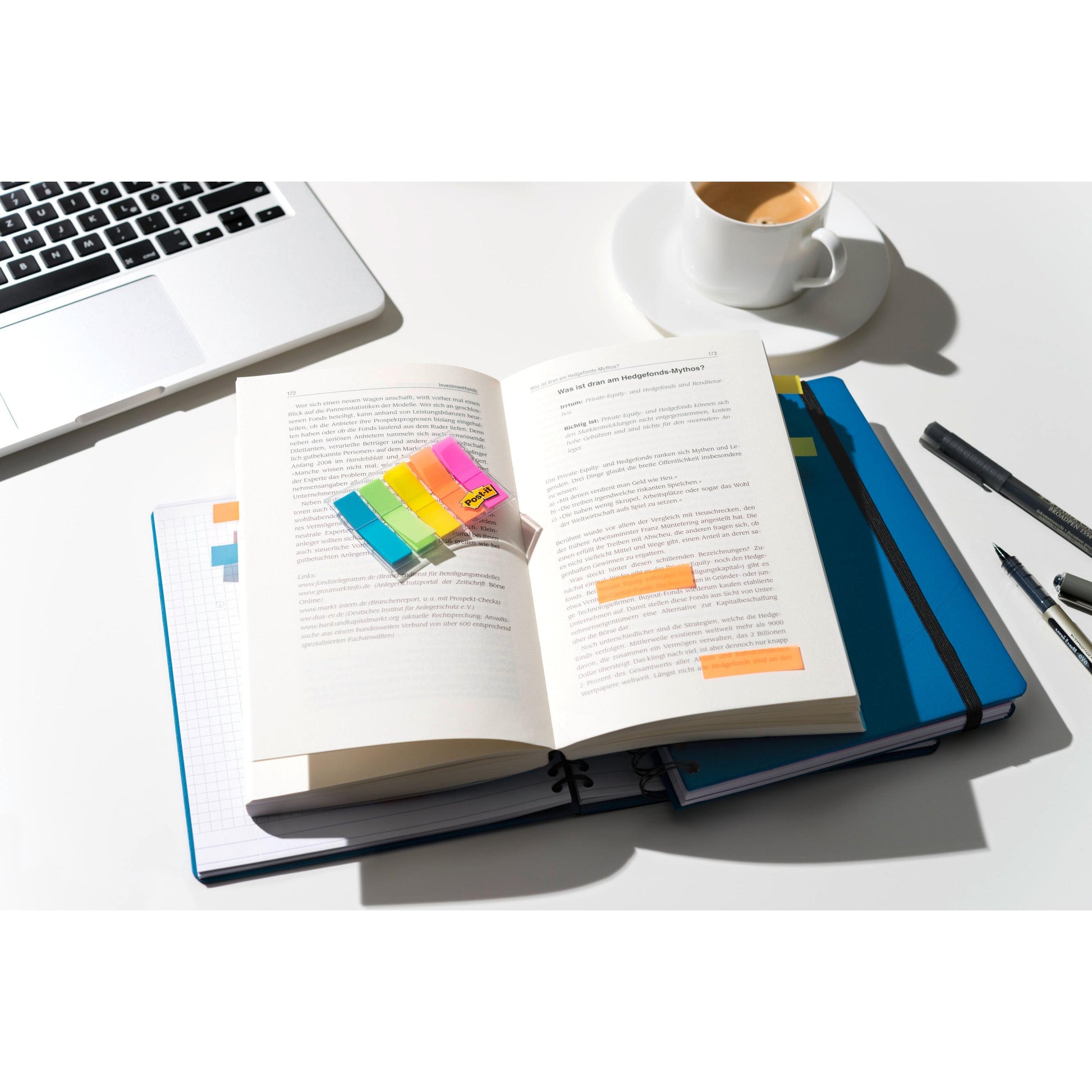 Auf einem gut beleuchteten Schreibtisch liegt ein aufgeschlagenes Buch, beschriftet mit 3M Deutschland GmbH Post-it® Index Haftstreifen Mini (11,9 x 43,2 mm, 100/Spender), über einem blauen Notebook und Laptop, daneben Stifte und Kaffee auf der weißen Fläche.