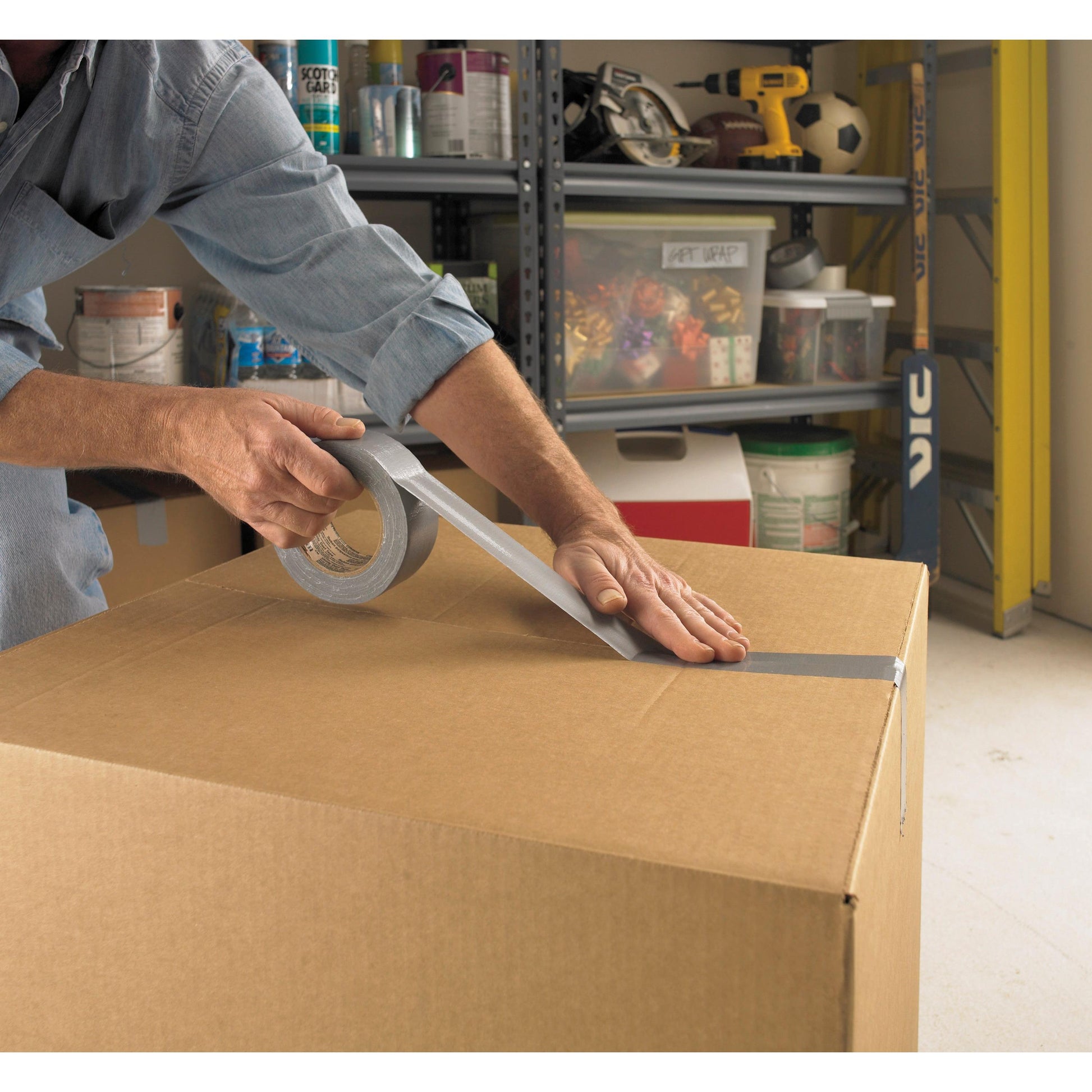 Eine Person verschließt einen großen Karton mit Scotch® Extremium™ Universal Klebeband (3 m x 19 mm) von 3M Deutschland GmbH in einer Garage. Im Hintergrund sind Regale mit Werkzeug, Farbdosen und anderen Gegenständen zu sehen.