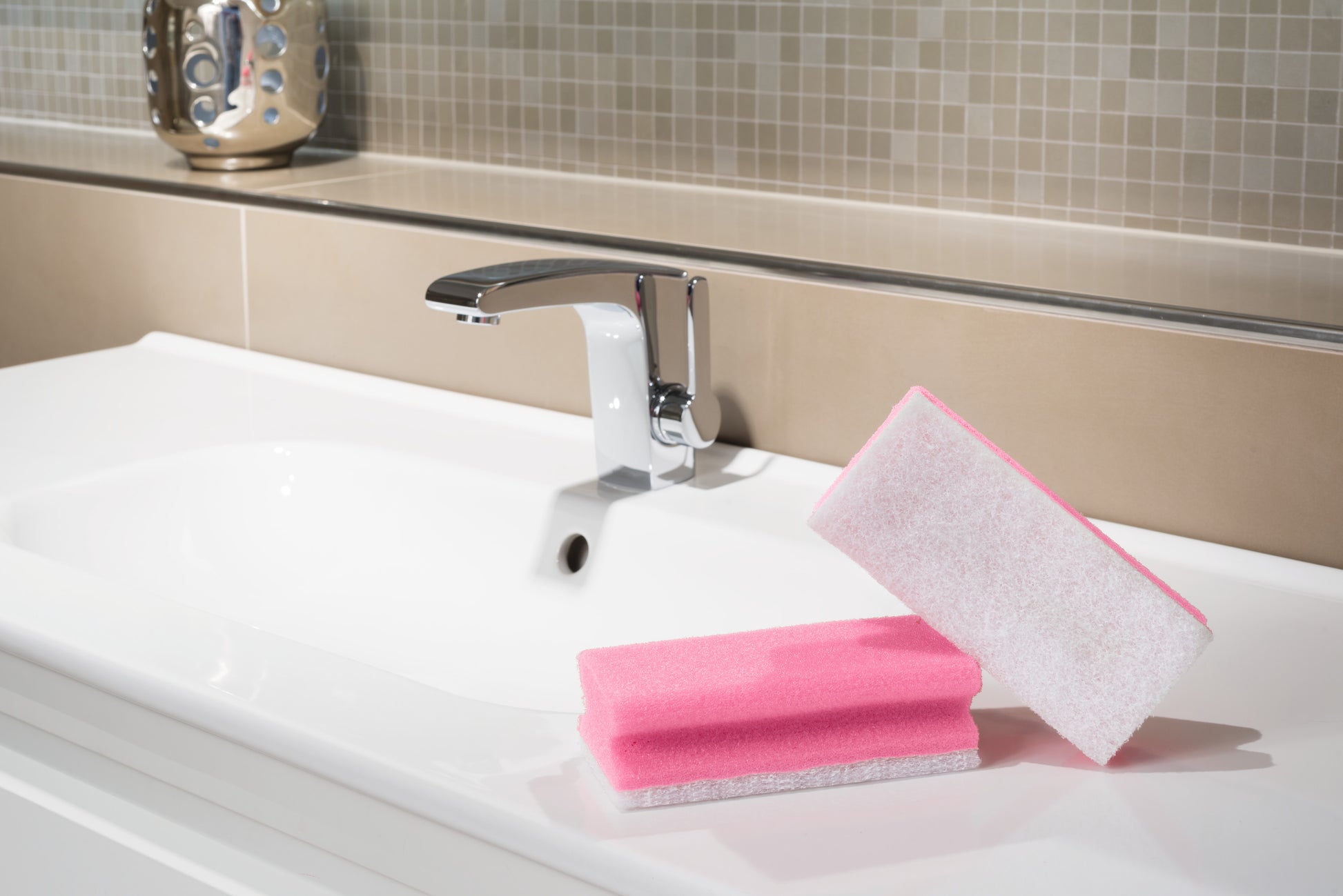 Abgebildet ist ein modernes Waschbecken mit verchromtem Wasserhahn. Auf der weißen Arbeitsplatte liegt ein Scotch-Brite® Bath Griffschwamm XXL der 3M Deutschland GmbH, während beige- und cremefarbene Mosaikfliesen die Aufkantung zieren.