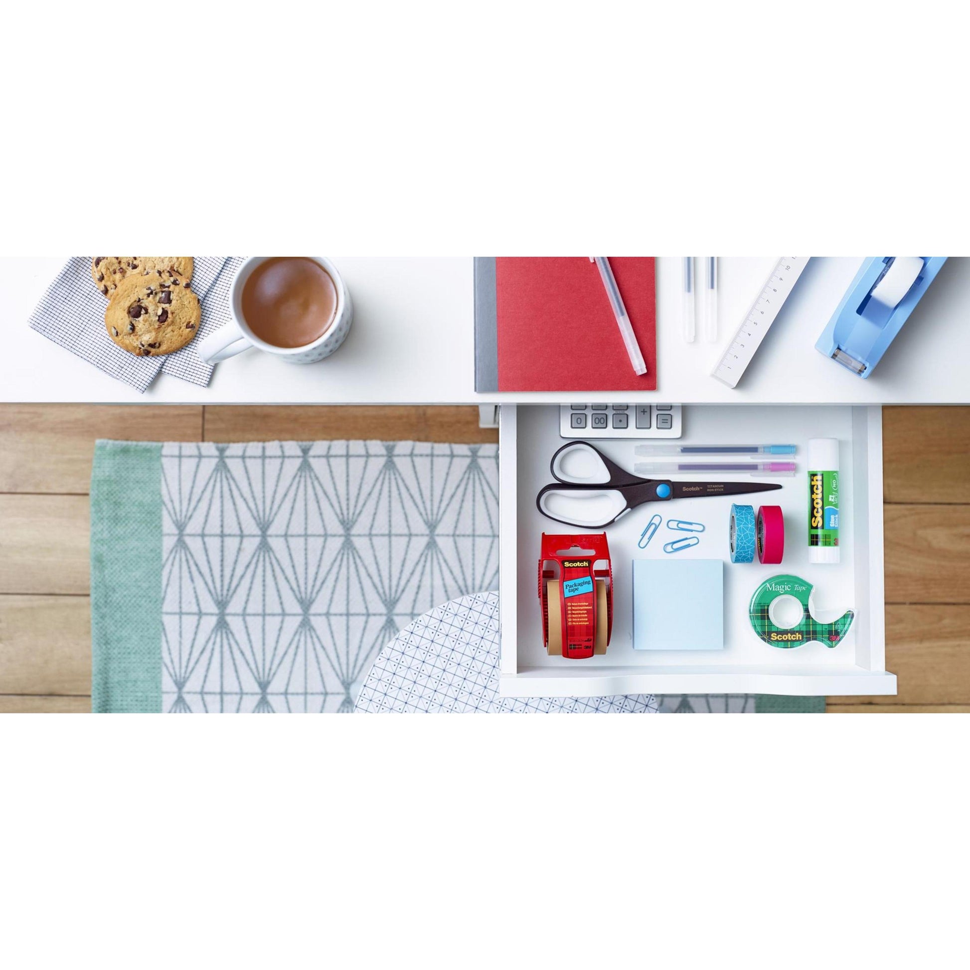 Offene Schreibtischschublade mit einer Scotch™ Titanschere, 20 cm von 3M Deutschland GmbH, Klebeband, Klebestift, Haftnotizen, Büroklammern und Korrekturband. Auf dem Schreibtisch: rotes Notizbuch, Stift, Lineal, Klebebandabroller, Kaffeetasse und Kekse auf einer Serviette.