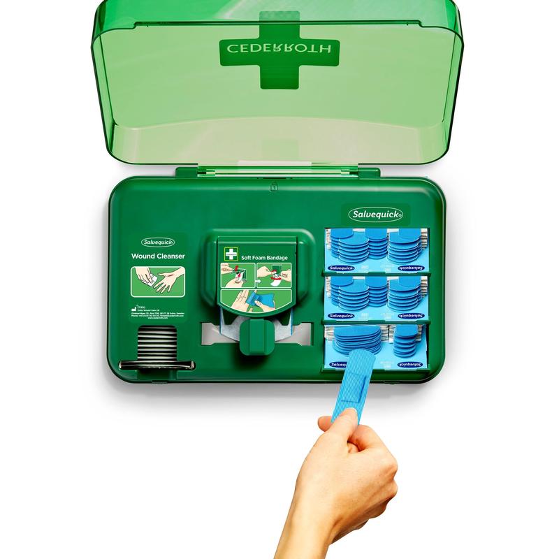 Ein geöffneter Wundversorgungsspender, blau (Orkla Wound Care AB) zeigt Wundreinigungstücher auf der linken Seite und blaue weiche Schaumstoffverbände auf der rechten Seite, während eine Hand ein blaues Werkzeug benutzt, um einen Verband aus dem Spenderteil zu ziehen.