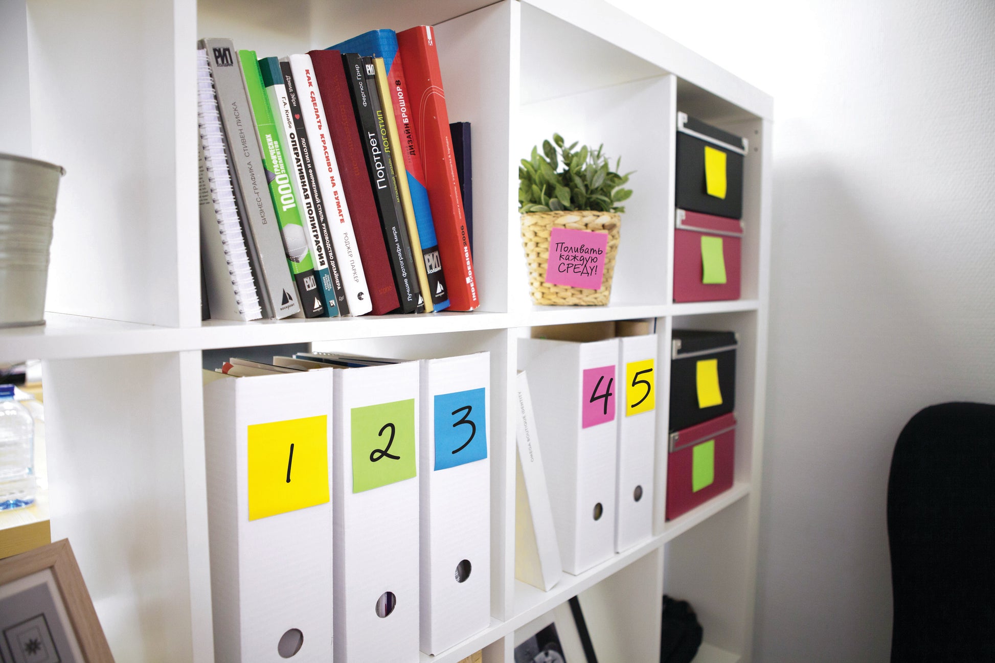 Ein weißes Regal enthält Bücher, Ordner mit den Nummern 1 bis 5 und Post-it®-Zettel in verschiedenen Neonfarben (3M Deutschland GmbH), rosa Aufbewahrungsboxen, eine Pflanze in einem gelben Topf und verschiedene Büromaterialien.