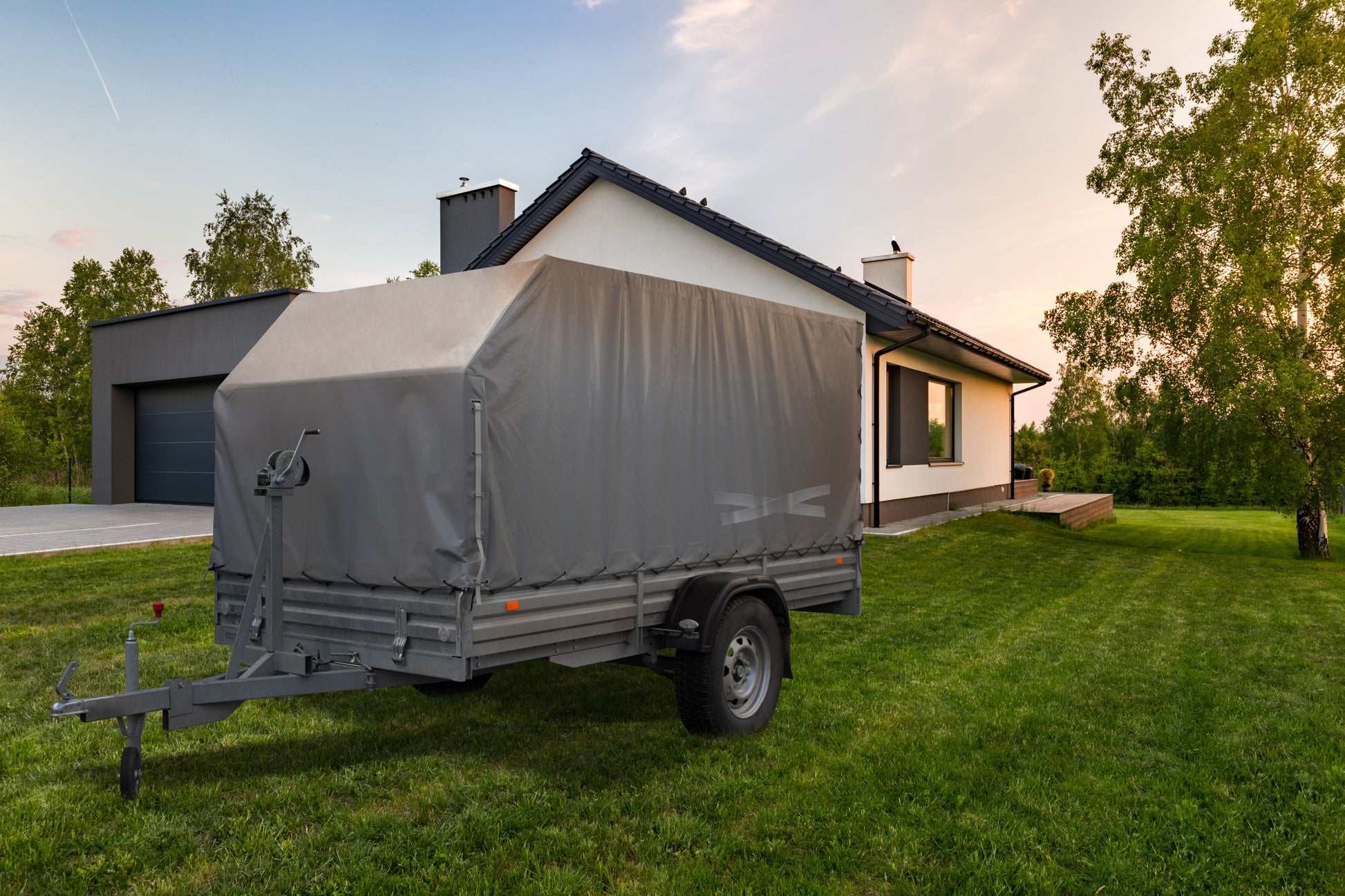 Ein grauer Nutzfahrzeuganhänger, der mit dem Scotch® Extremium™ All Weather Hochleistungs-Klebeband (27m x 48mm) der 3M Deutschland GmbH gesichert ist, steht bei teilweise bewölktem Himmel auf einer Wiese vor einem modernen weißen Haus mit dunklem Dach und angeschlossener Garage.