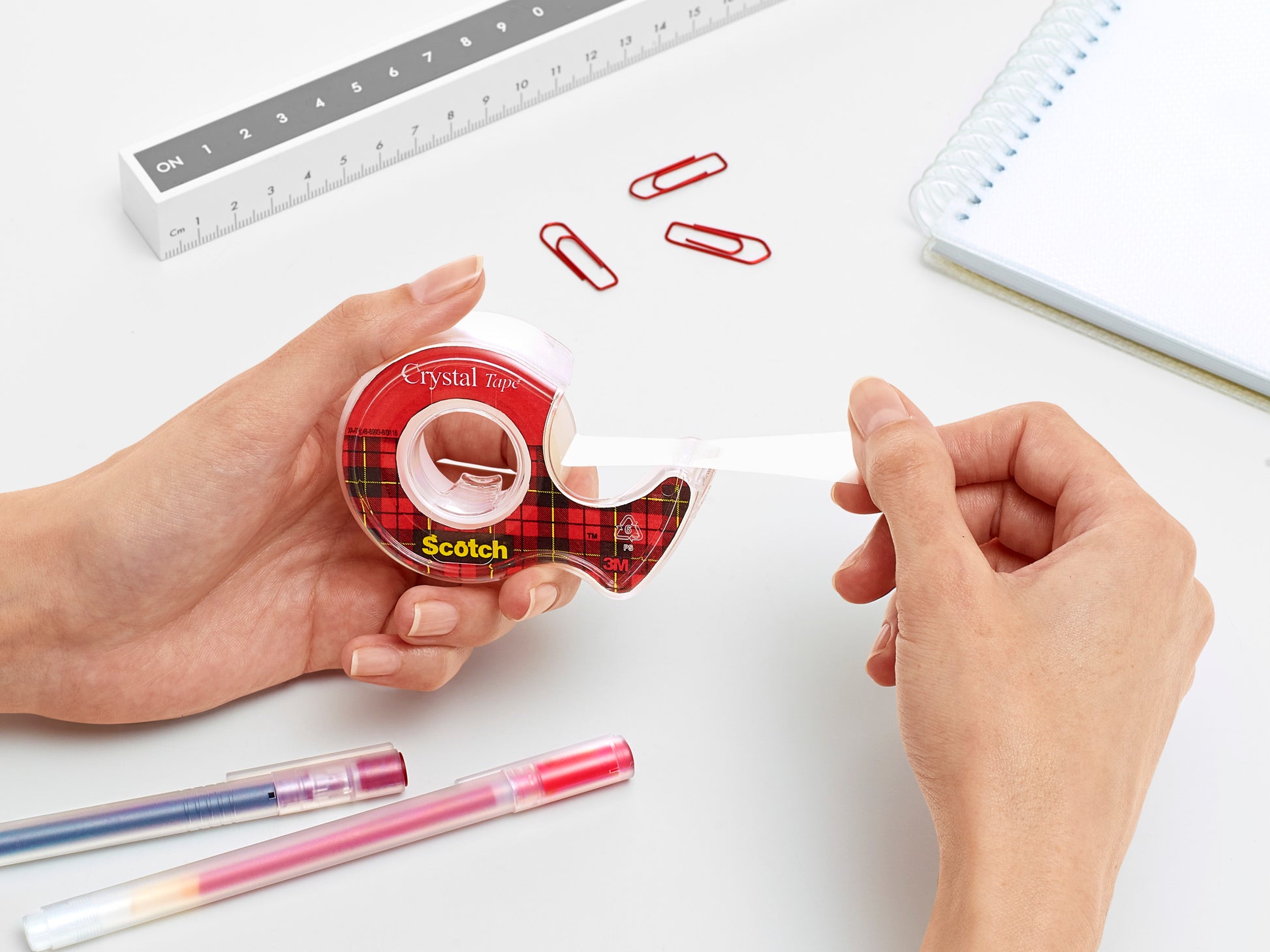 Eine Person zieht Scotch® Crystal Klebeband (19 mm x 7,5 m) aus dem Handabroller der 3M Deutschland GmbH. Auf dem weißen Schreibtisch liegen rote Büroklammern, ein Lineal, ein Notizbuch und zwei farbige Stifte.