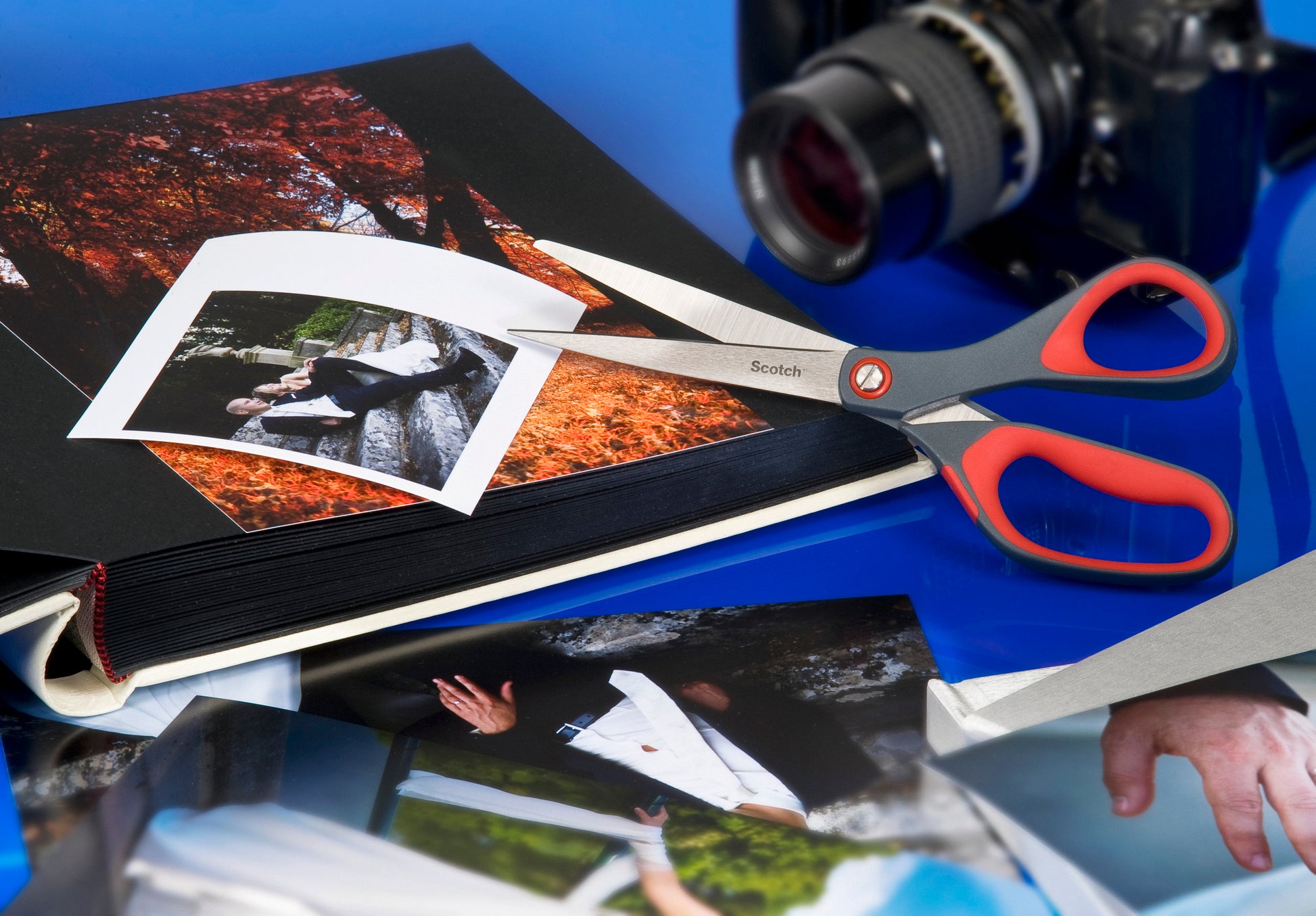 Eine Scotch™ Präzisionsschere rot von 3M Deutschland GmbH liegt auf einem geöffneten Fotoalbum, eine lose Fotografie ist teilweise geschnitten. Daneben befinden sich weitere Fotos und eine Kamera auf einer blauen Fläche.