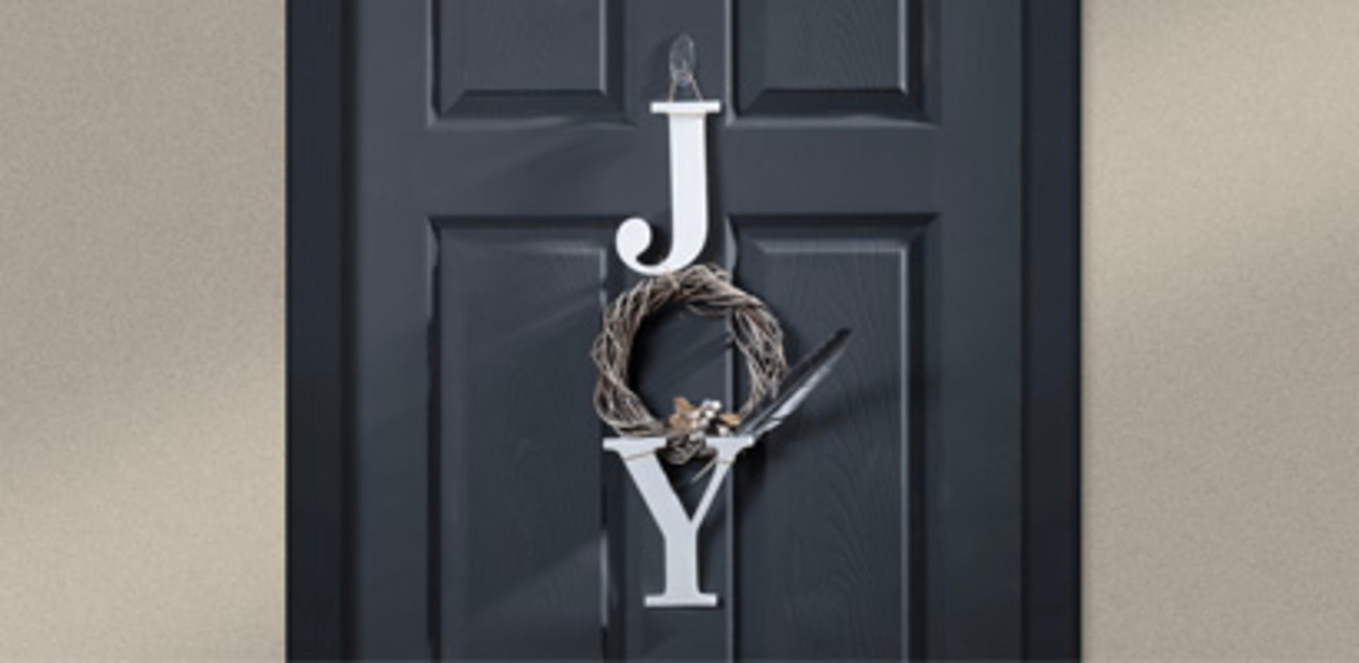 An einer schwarzen Tür steht "JOY" senkrecht: Das "J" und das "Y" sind weiß, während das "O" ein Kranz mit einer kleinen Schleife ist, der mit einem 3M Command™ transparenten, ovalen Outdoor-Haken (17091AW, 1 Stück) der 3M Deutschland GmbH aufgehängt wurde.