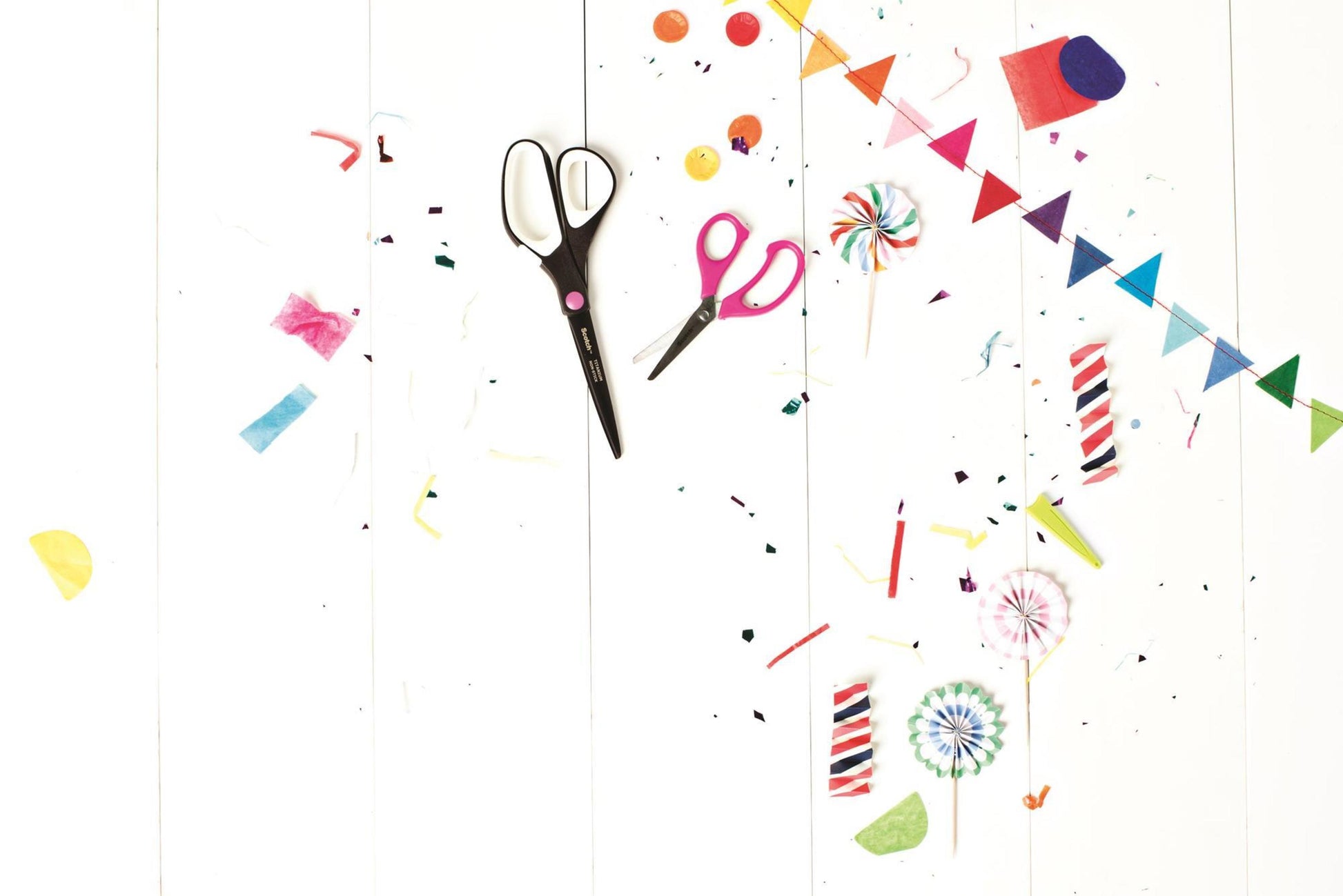 Eine Scotch™ Titanschere (20 cm) von 3M Deutschland GmbH, bunte Papierschnipsel, Konfetti, Lutscher, Kerzen und ein kleines Regenbogenbanner sind auf einer weißen Holzfläche verstreut und sorgen für eine festliche, kreative Stimmung.