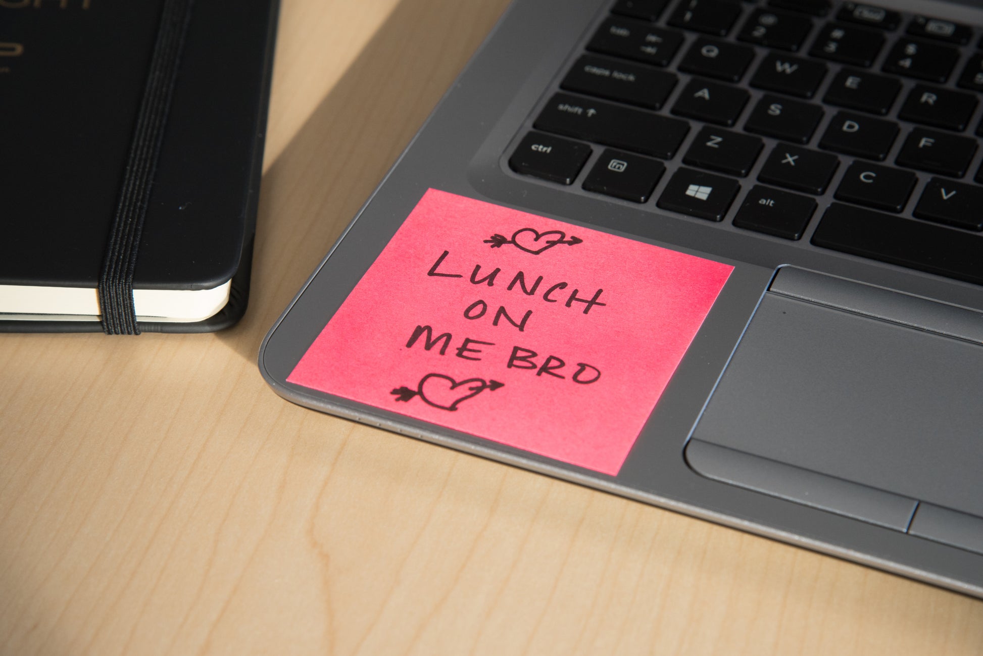Ein Post-it® Notes Würfel (76x76mm, 450 Blatt, 100% PEFC) von 3M Deutschland GmbH in Pink sitzt auf einer Laptop-Tastatur mit "LUNCH ON ME BRO" und Herz-Kritzeleien. Ein geschlossenes schwarzes Notizbuch liegt daneben auf einem hellen Holztisch.