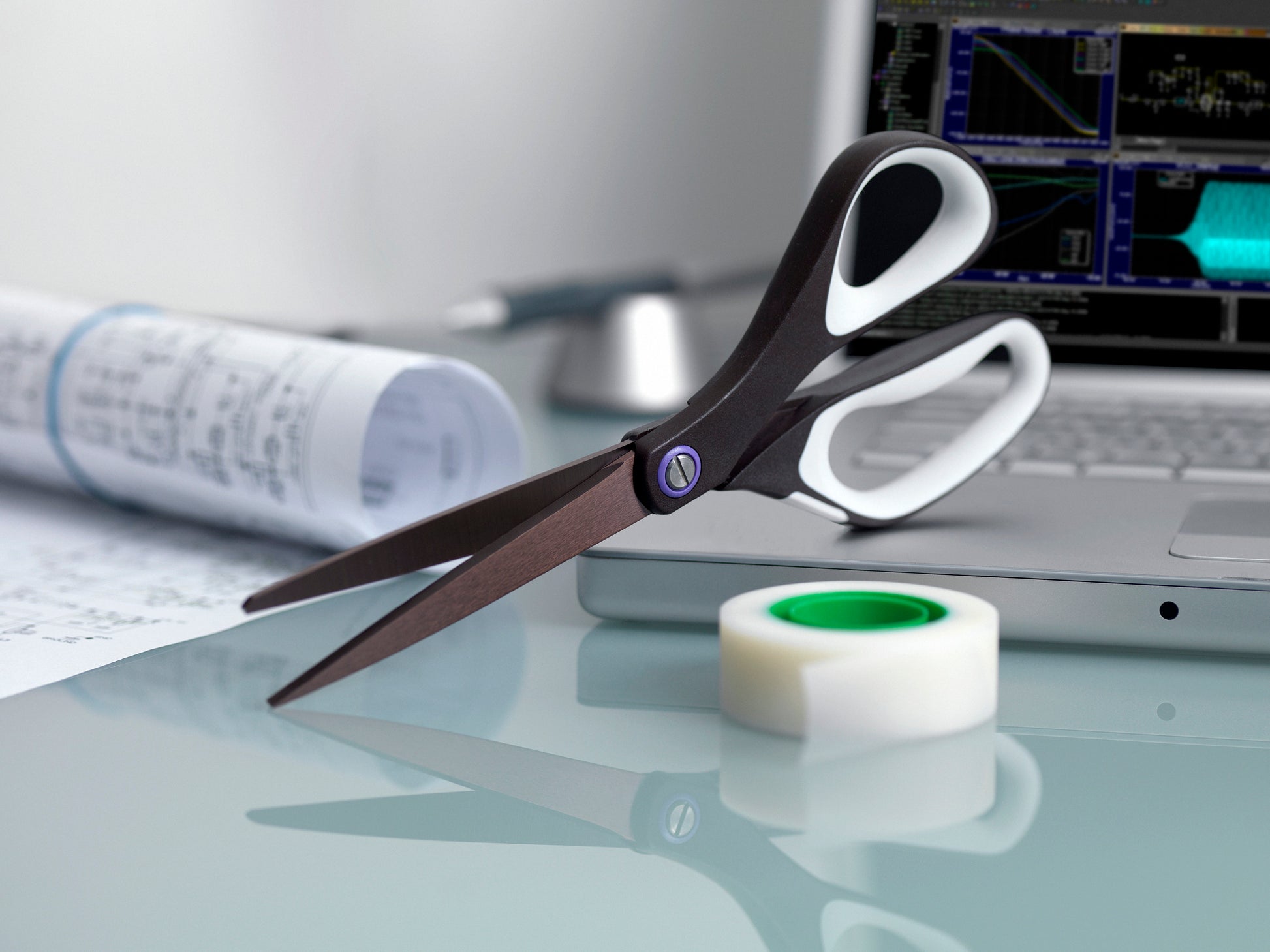 Eine 3M Deutschland GmbH Scotch™ Titanschere, 20 cm, eine Rolle durchsichtiges Klebeband, Dokumente und ein Laptop mit Diagrammen auf dem Bildschirm sind auf einem Glastisch platziert.