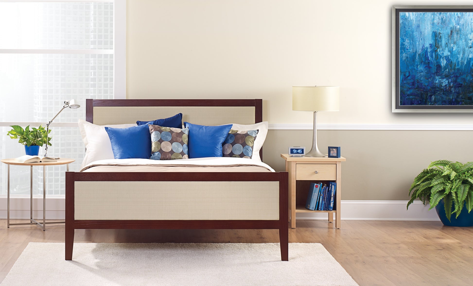 Ein ordentlich gemachtes Bett mit weißen und blauen Kissen steht vor einer hellen Wand, die mit ScotchBlue™ Malerabdeckband "Multi-Surface" der 3M Deutschland GmbH beklebt ist. Auf einem Nachttisch stehen eine Lampe, eine Uhr, Bücher und eine Topfpflanze neben dem hellen Fenster.