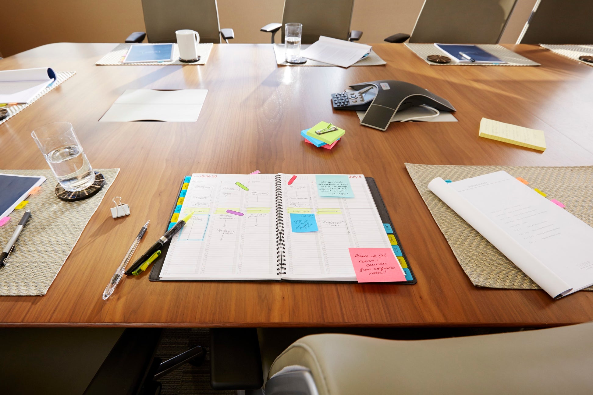 Ein Besprechungstisch mit einem offenen Planer, Stiften, Papieren, einem Glas Wasser, einem Konferenztelefon und dem 3M Post-it® Z-Notes Dispenser Katze (weiß) mit pinkfarbenen Super Sticky Z-Notes (76x76mm), 90 Blatt/Block. Leere Stühle umgeben den Tisch.