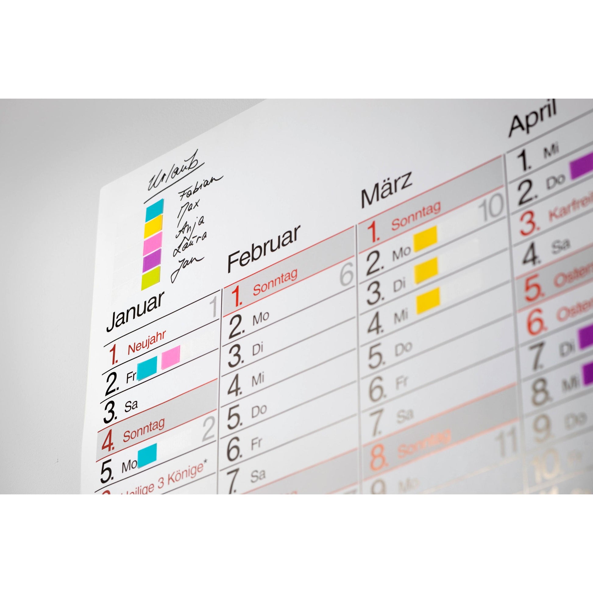 Nahaufnahme eines Wandkalenders für Januar bis März mit bunten 3M Deutschland GmbH Post-it® Index Haftstreifen (Mini, 11,9 x 43,2 mm) als farbcodierte Marker für die handschriftlichen Termine oder Aktivitäten jeder Person.