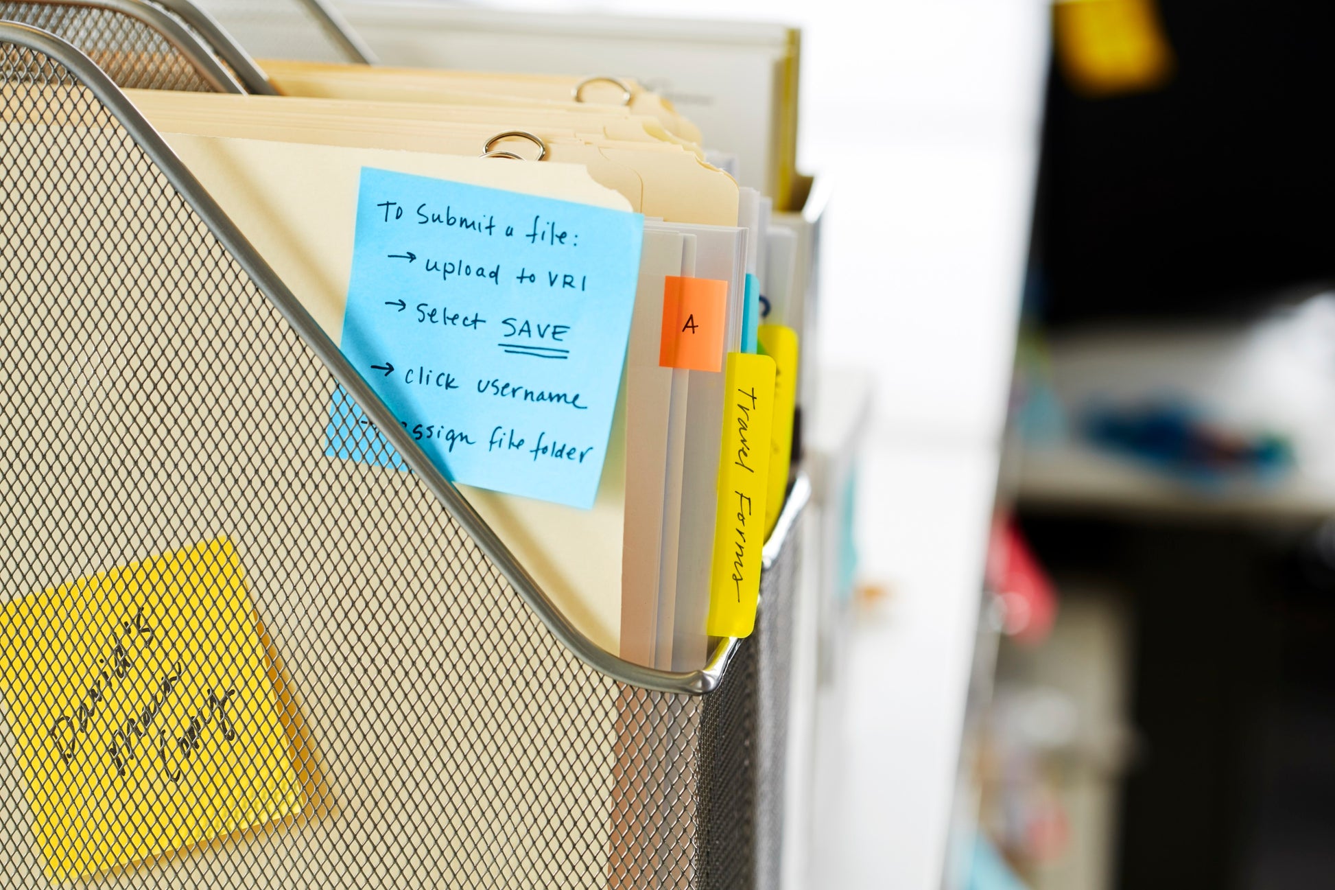 Ein Drahtgeflecht-Ordner hält Manila-Ordner mit farbigen Reitern und handgeschriebenen Etiketten. Ein blauer Post-it® Notes Würfel (76x76 mm, 450 Blatt/Block, 100% PEFC) von 3M Deutschland GmbH ist angebracht; weitere Haftnotizen erscheinen im Hintergrund.