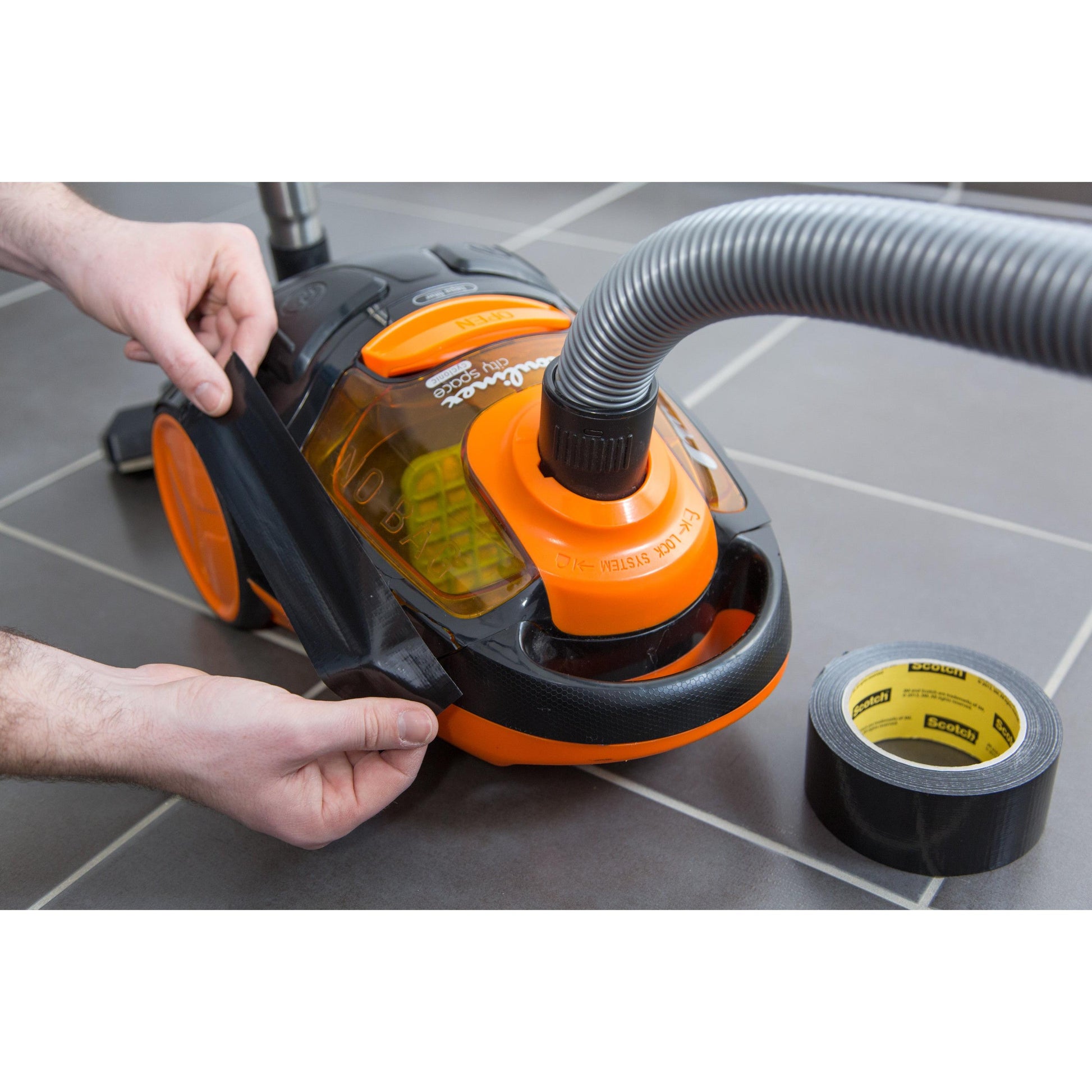 Eine Person repariert das rissige orange-schwarze Gehäuse eines Staubsaugers mit Scotch® Extremium™ Universal Klebeband (3 m x 19 mm) der 3M Deutschland GmbH, wobei die Rolle auf dem grauen Fliesenboden in der Nähe liegt.