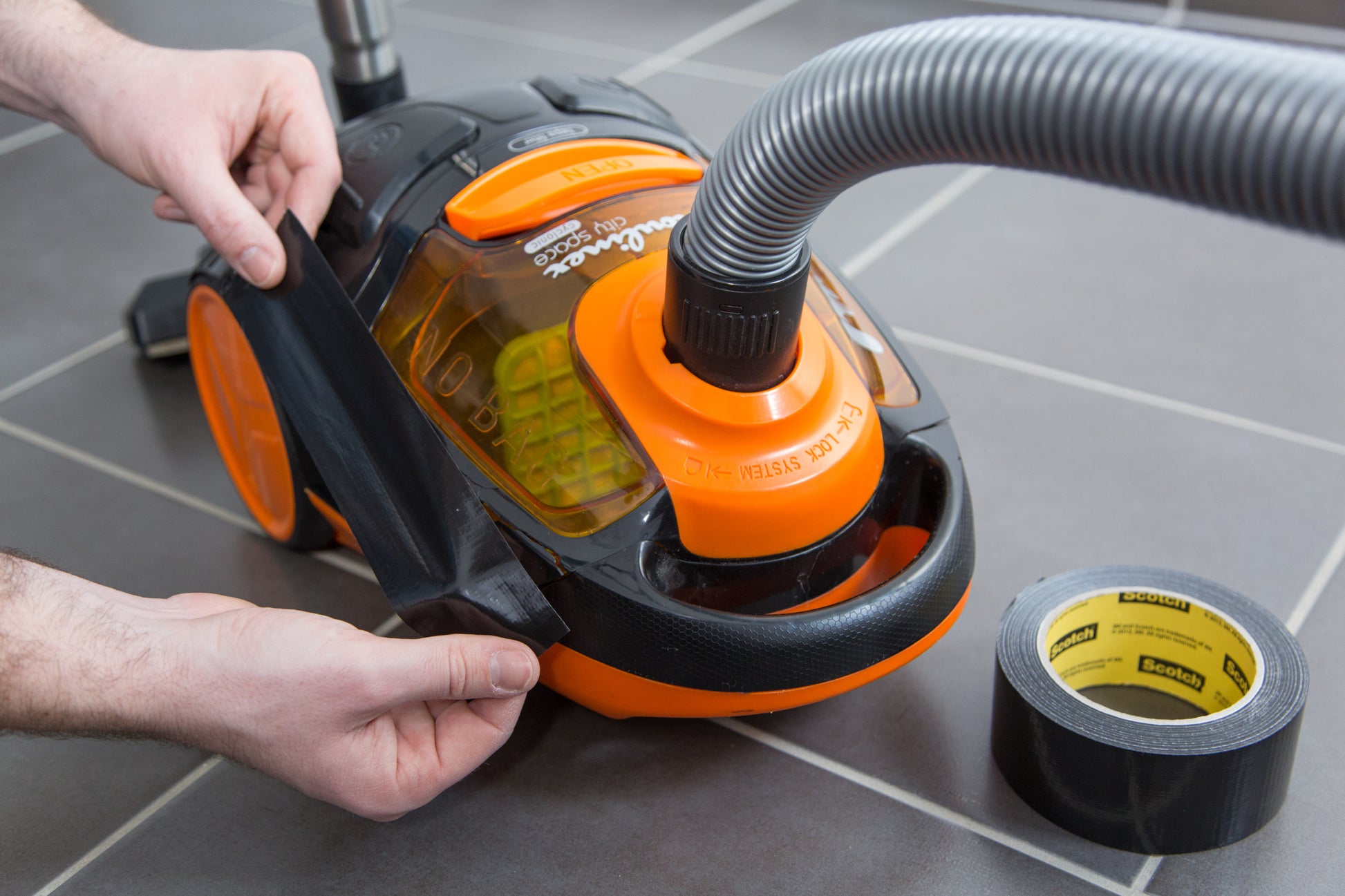 Eine Person klebt Scotch® Universal Klebeband 2904 von 3M Deutschland GmbH auf die Seite eines orange-schwarzen Staubsaugers auf einem grauen Fliesenboden, die Klebebandrolle liegt daneben.