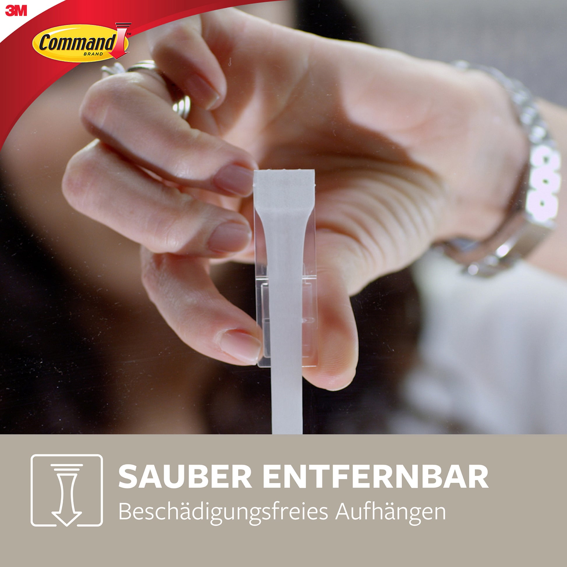 Eine Hand zieht eine Lasche an einem transparenten Klebestreifen, der an einem Glas befestigt ist, und zeigt das 3M Command-Logo und deutsche Informationen über den Mittelgroßen Command™ Metallhaken in gebürstetem Nickel (1 Haken, 2 Streifen) von 3M Deutschland GmbH.