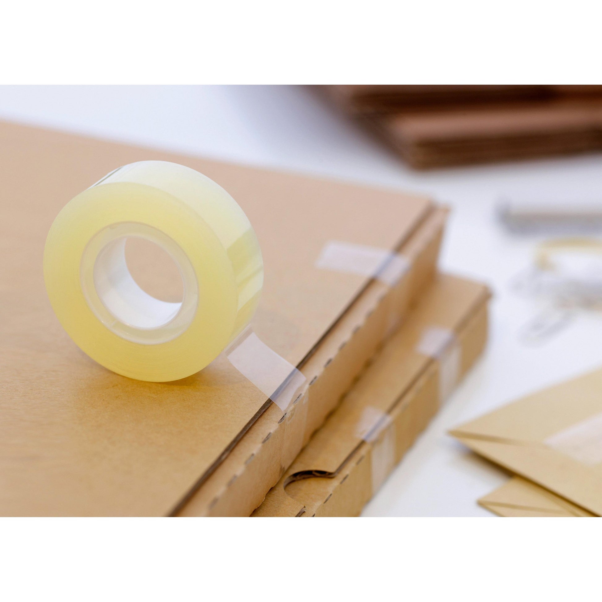 Eine Rolle Scotch® Transparentes Klebeband 550 der 3M Deutschland GmbH liegt auf Kartons, die mit dem Klebeband gesichert sind. Im Hintergrund sind Umschläge und andere Verpackungsmaterialien zu sehen.