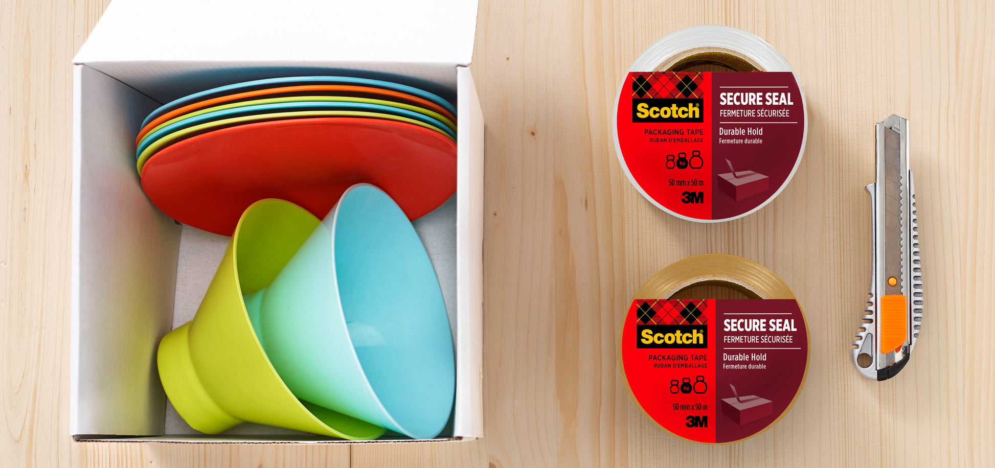 Eine Pappschachtel mit bunten Tellern und Schalen steht auf einer Holzfläche neben zwei Rollen Scotch® Verpackungsklebeband für einen sicheren Verschluss, 50 mm x 66 m von 3M Deutschland GmbH und einem Universalmesser.