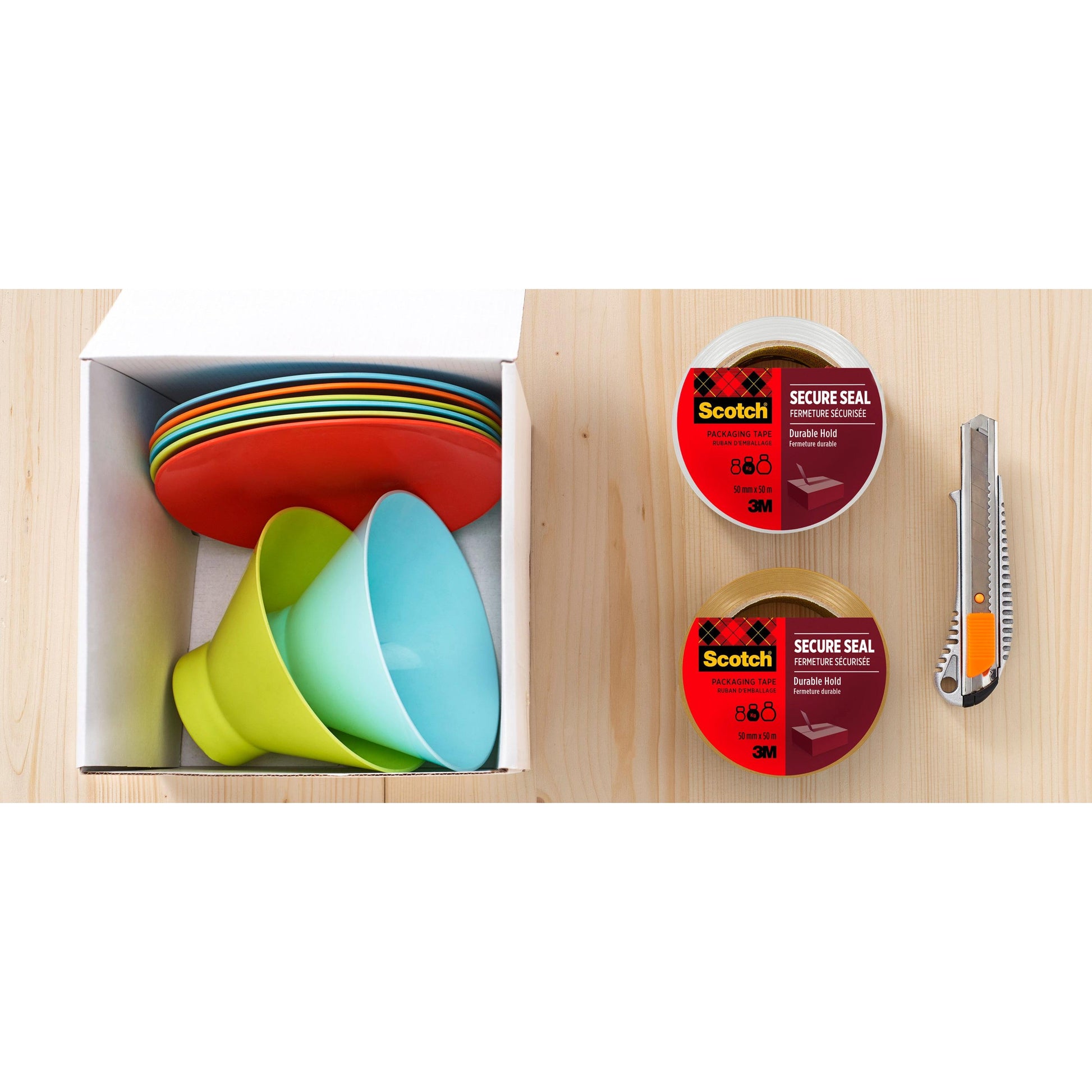 Eine Pappschachtel mit bunten Tellern und Schalen steht auf einer Holzfläche neben zwei Rollen Scotch® Verpackungsklebeband für einen sicheren Verschluss, 50 mm x 66 m von 3M Deutschland GmbH und einem Universalmesser.