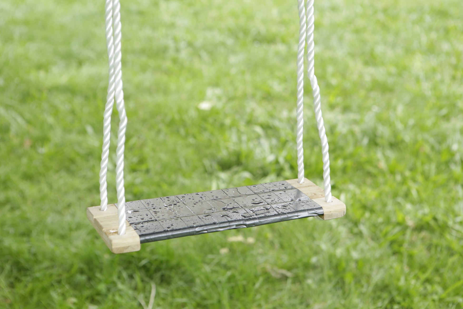 Nahaufnahme einer einfachen Schaukel mit Holzsitz und weißen Seilen, gesichert mit Scotch® Extremium™ All Weather Hochleistungs-Klebeband (27 m x 48 mm) von 3M Deutschland GmbH, die über grünem Gras mit Wassertropfen hängt.