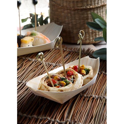 Drei gerollte Tortilla-Schnecken, gefüllt mit Gemüse und mit Holzspießen befestigt, werden in einer „pure“ Holz Fingerfood-Schale (Schiffchen) der PAPSTAR GmbH auf einer gewebten Unterlage serviert, im Hintergrund ist verschiedenes Fingerfood zu sehen.