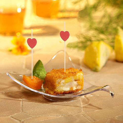 In einer kleinen Glasschale befindet sich ein panierter, frittierter Würfel mit weißer Füllung, garniert mit einem Blatt und einer Orange. Zwei PAPSTAR 100 Fingerfood - Picker der PAPSTAR GmbH, garniert mit roten Herzen, sind enthalten. Im Hintergrund sind Zitronenspalten zu sehen.