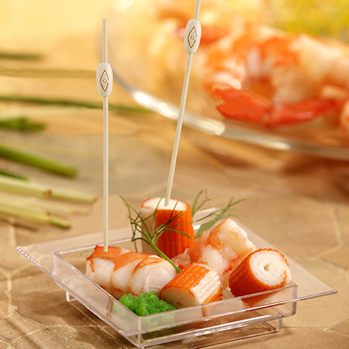 Eine quadratische, durchsichtige Schale enthält Krabbenimitate, Garnelen und grünen Rogen mit Kräutern. Zwei PAPSTAR 100 Fingerfood - Picker der PAPSTAR GmbH sind in die Meeresfrüchte eingesetzt. Im Hintergrund erscheinen unscharfe Garnelen.