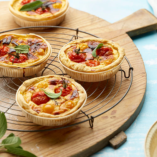 Drei kleine Gemüsequiches mit Kirschtomaten und Kräutern liegen auf einem runden Drahtgestell auf einem Holzbrett, umgeben von frischen Kräuterblättern und PAPSTAR 50 Backformen "pure" braun der PAPSTAR GmbH.