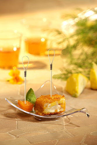Nahaufnahme eines Vorspeisentellers mit zwei mundgerechten Snacks auf PAPSTAR 100 Fingerfood - Picker von PAPSTAR GmbH, hergestellt aus Bambus Holz und garniert mit Früchten, auf einer beigen Fläche mit Getränken und Kräutern im Hintergrund.