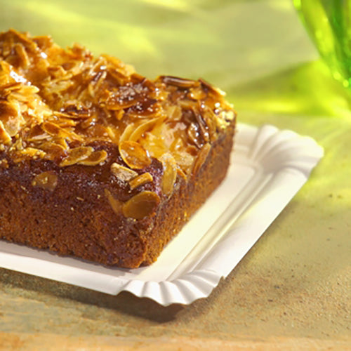 Ein Stück Mandelkuchen liegt auf einem weißen, rechteckigen PAPSTAR „pure“ Pappteller (13x20 cm) der PAPSTAR GmbH. Im Hintergrund ist ein grünes Glas unscharf zu erkennen.