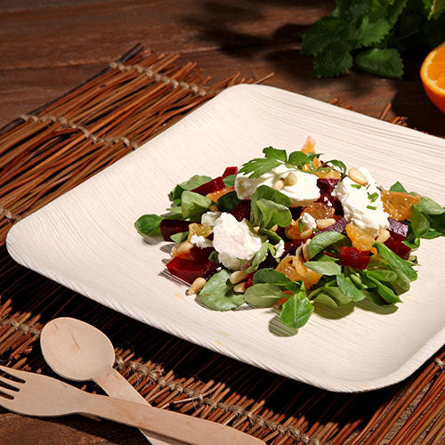 Ein PAPSTAR 6 Teller, Palmblatt „pure“ eckig der PAPSTAR GmbH, enthält einen gemischten grünen Salat mit Roter Bete, Orangenscheiben, Ziegenkäse und Nüssen. Er steht auf einer Bambusmatte mit Holzbesteck; im Hintergrund sind eine Orange und Kräuter zu sehen.