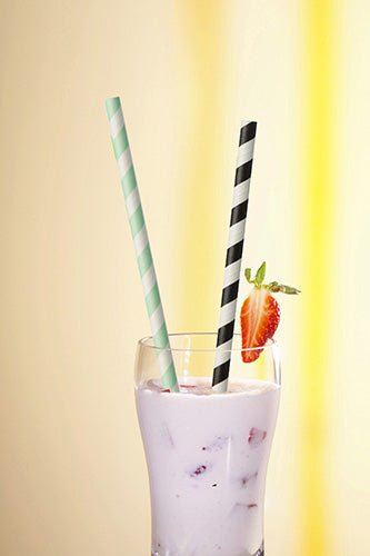 In einem Glas mit rosa Milchshake, Eis, Fruchtstückchen und einer Erdbeergarnitur befinden sich zwei PAPSTAR 100 Trinkhalme Papier Ø 6 mm (by PAPSTAR GmbH), ein grün gestreiftes und ein schwarz gestreiftes.