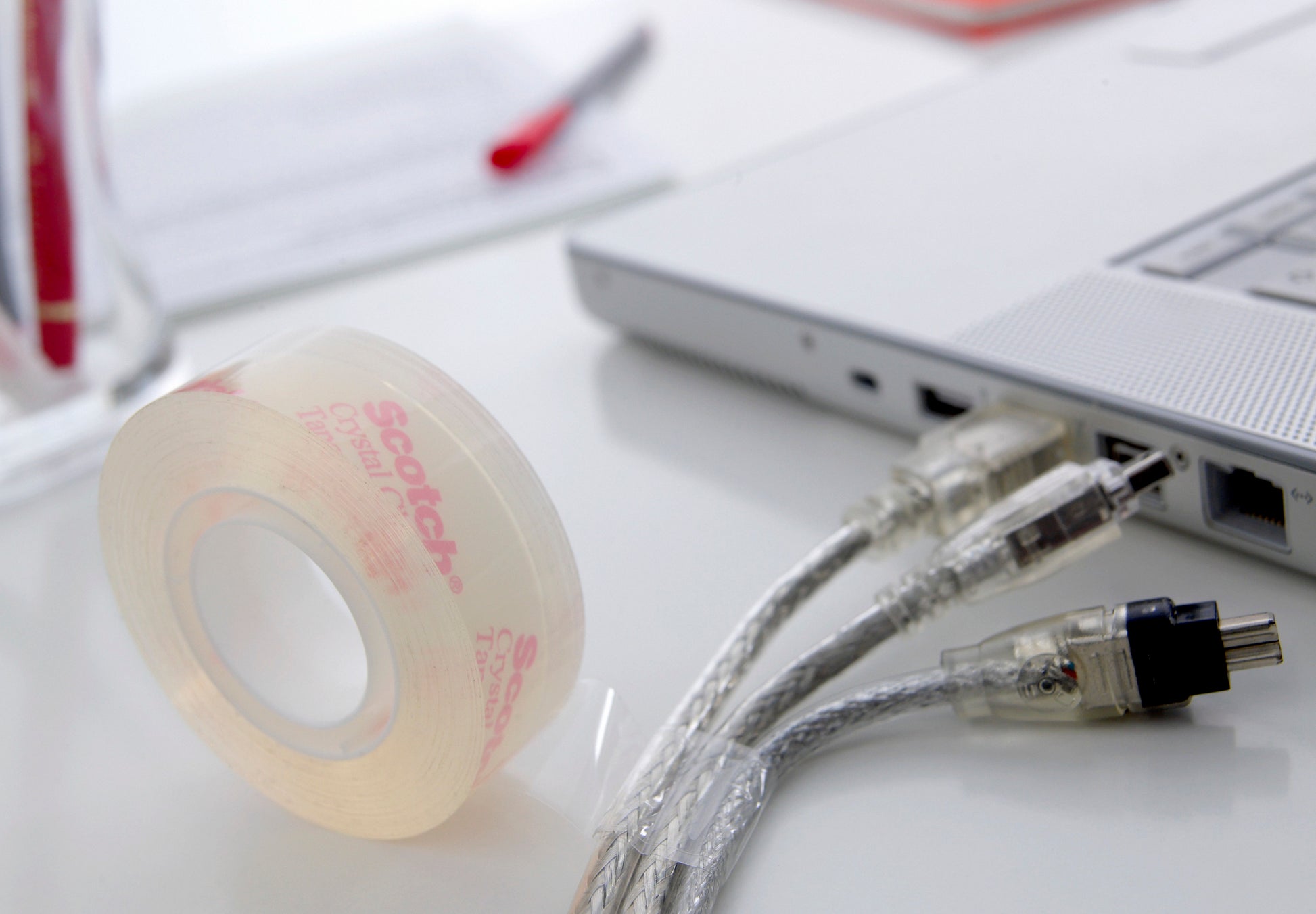 Eine Rolle Scotch® Crystal Klebeband der 3M Deutschland GmbH (19 mm x 33 m, Vorteilspack, 14 Rollen) liegt auf einem weißen Schreibtisch neben einem Laptop mit zwei eingesteckten durchsichtigen Kabeln; ein roter Stift und Papiere sind unscharf im Hintergrund zu sehen.