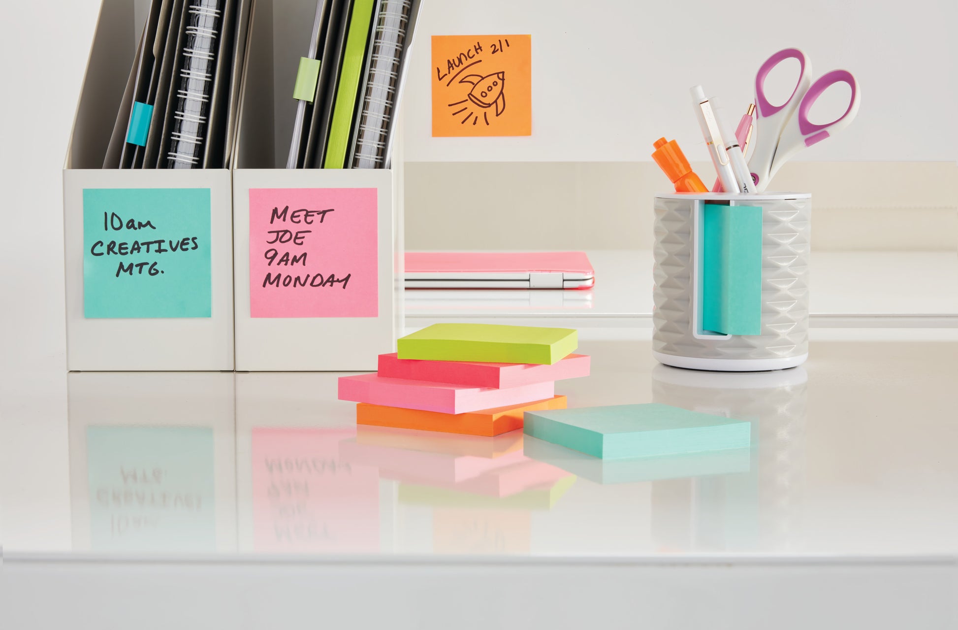 Ein aufgeräumter Schreibtisch mit den farbenfrohen Post-it® Notes Würfel (76x76mm, 450 Blatt/Block, 100% PEFC) der 3M Deutschland GmbH, einem weißen Stifteköcher, gefüllt mit Stiften und Scheren, Notizbüchern und Karteikästen. Die Haftnotizen sind mit handschriftlichen Erinnerungen versehen.