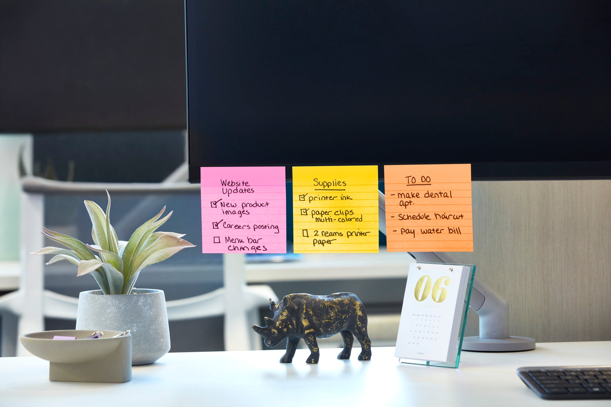 Ein Computermonitor zeigt bunte Post-it® Super Sticky Notes im Großformat (101 mm x 101 mm, 90 Blatt/Block, 100% PEFC) von 3M Deutschland GmbH mit Website-Updates, Material und Aufgaben. Auf dem Schreibtisch stehen eine Pflanze, eine schwarze Elefantenfigur und ein Tischkalender mit der Jahreszahl "06".