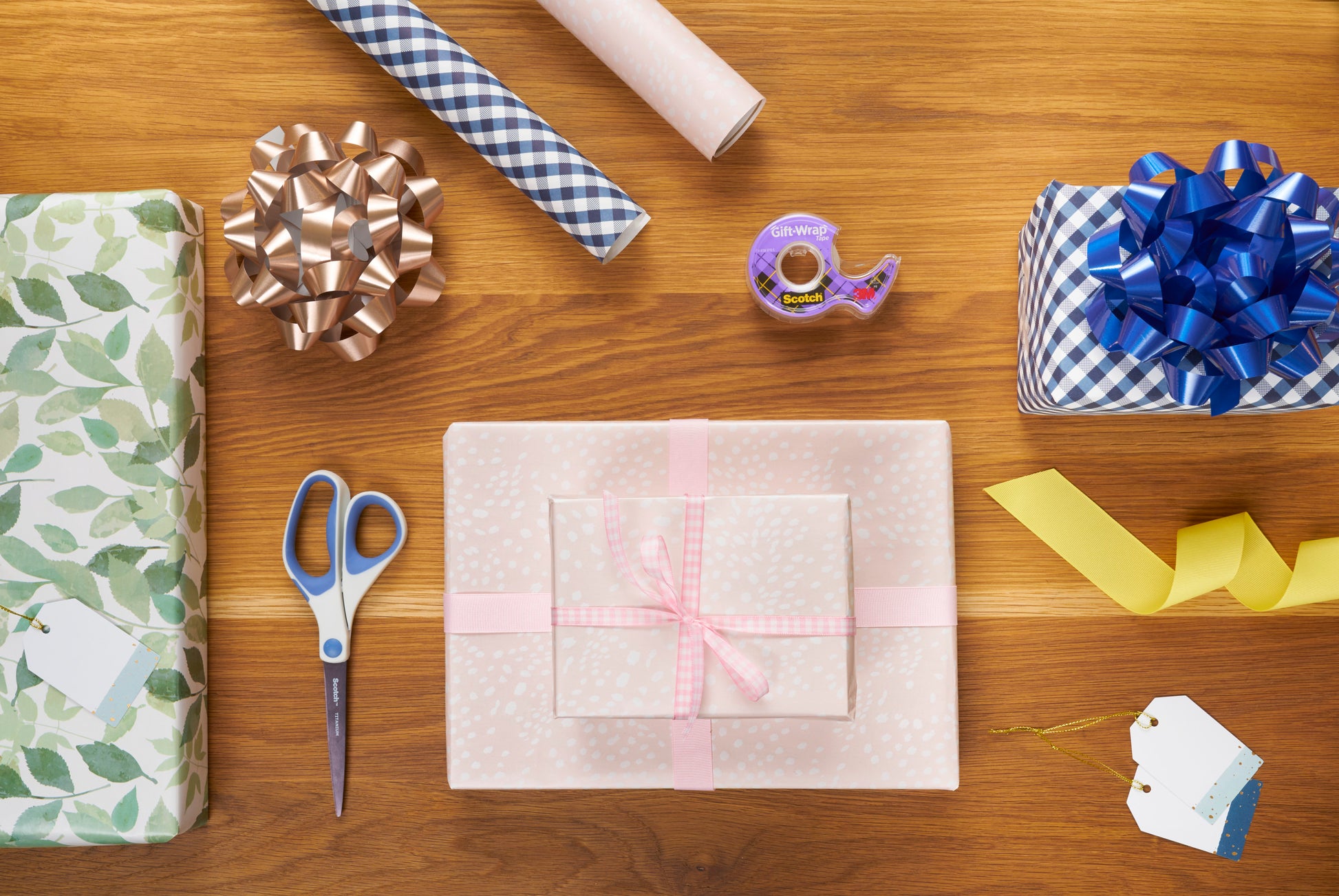 Ein Holztisch mit eingepackten Geschenken, Geschenkanhängern, Schleifen, Bändern, Scheren und einer Rolle Scotch® Geschenk-Satinklebeband von 3M Deutschland GmbH (19 mm x 25 m), alles fein säuberlich angeordnet zum Einpacken von Geschenken.