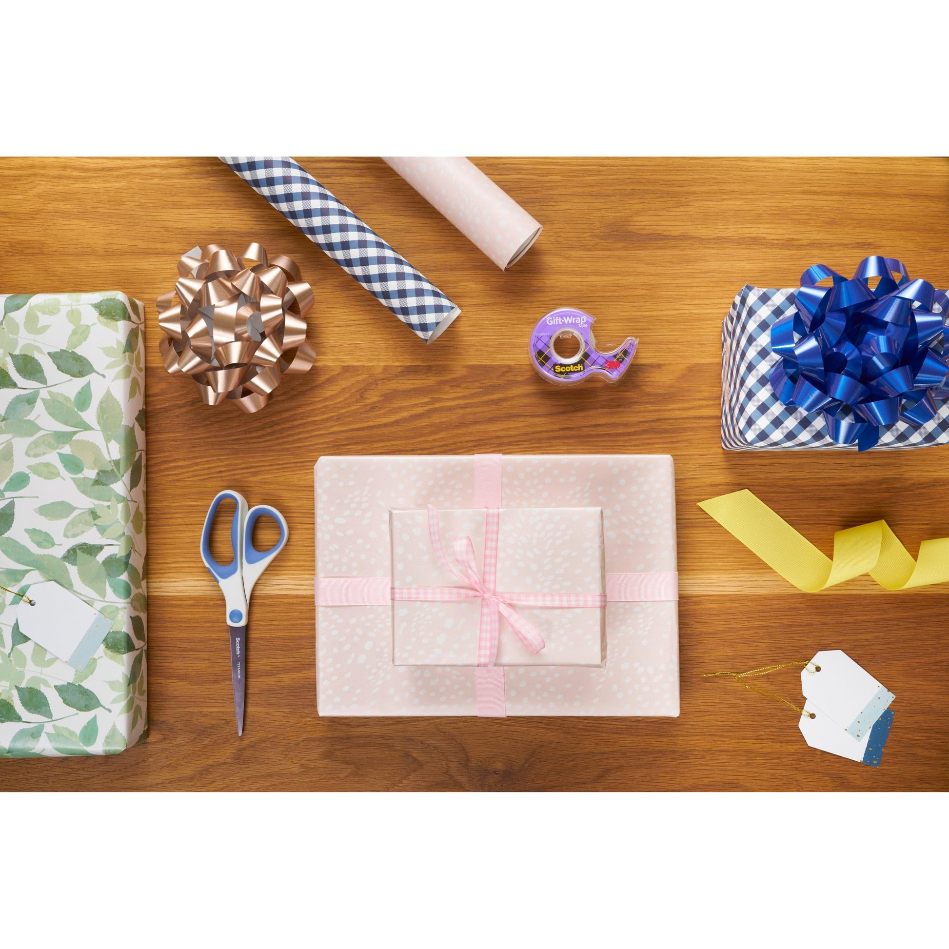 Ein Holztisch mit eingepackten Geschenken, Geschenkanhängern, Schleifen, Bändern, Scheren und einer Rolle Scotch® Geschenk-Satinklebeband von 3M Deutschland GmbH (19 mm x 25 m), alles fein säuberlich angeordnet zum Einpacken von Geschenken.