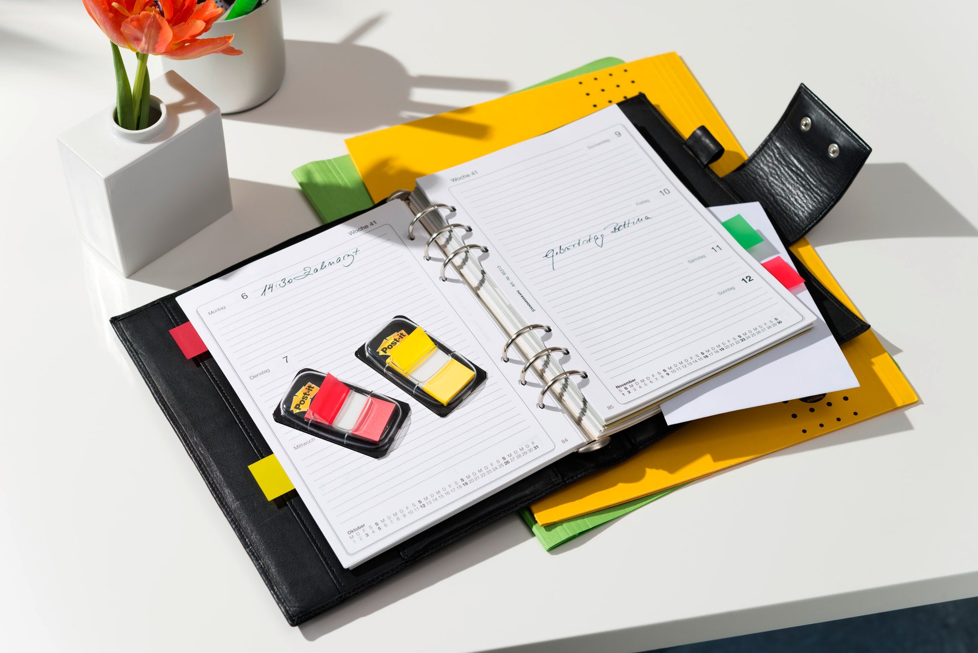 Ein ledergebundener Planer liegt offen auf einem Schreibtisch mit zwei Post-it® Index Haftstreifen (25,4 x 43,2 mm, verschiedene Farben), einem Stift, Dokumentenorganisationsmappen und einer Topfblume daneben.