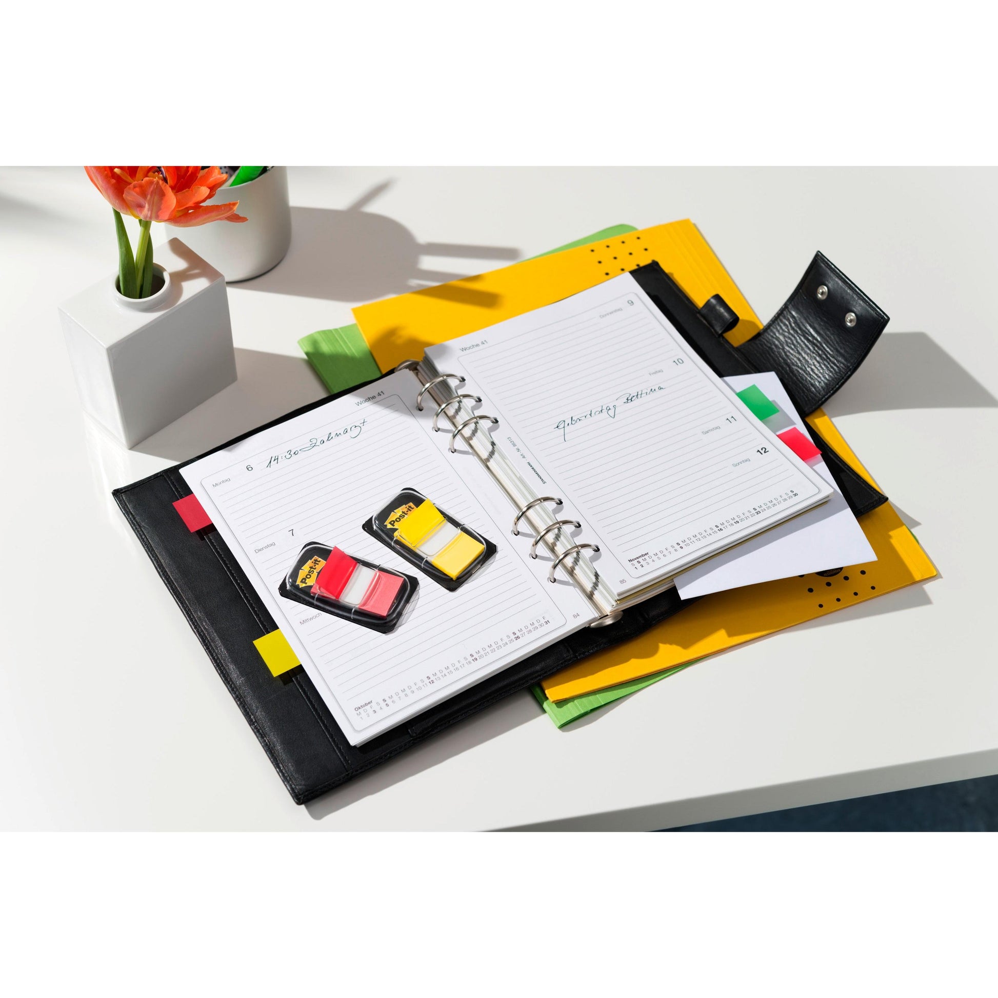 Ein schwarzer Ringbuchplaner aus Leder liegt aufgeklappt auf einem weißen Schreibtisch und zeigt 3M Deutschland GmbH Post-it® Index Haftstreifen (25,4 mm x 43,2 mm, 50/Stück) in Gelb und Rot. Daneben liegen Mappen in Gelb und Grün, lose Papiere und eine orangefarbene Kunstblume.