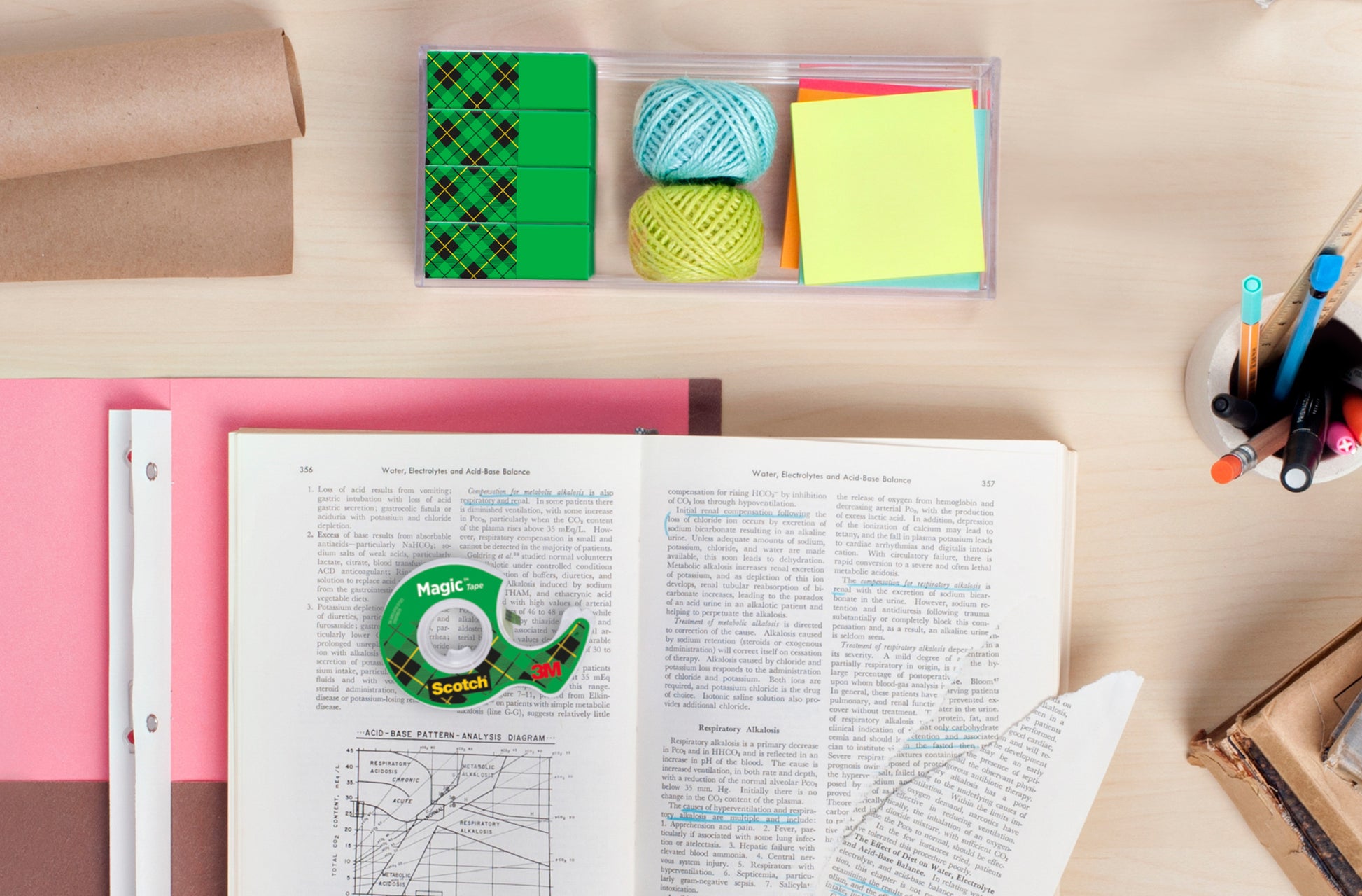 Ein Schreibtisch mit aufgeschlagenen Büchern, einer zerrissenen Seite, farbigen Haftnotizen, Radiergummis, Stiften, Bleistiften, Papier, Garnkugeln, einer Pappröhre und Scotch® Magic™ Unsichtbares Klebeband Vorteilspack von 3M Deutschland GmbH.