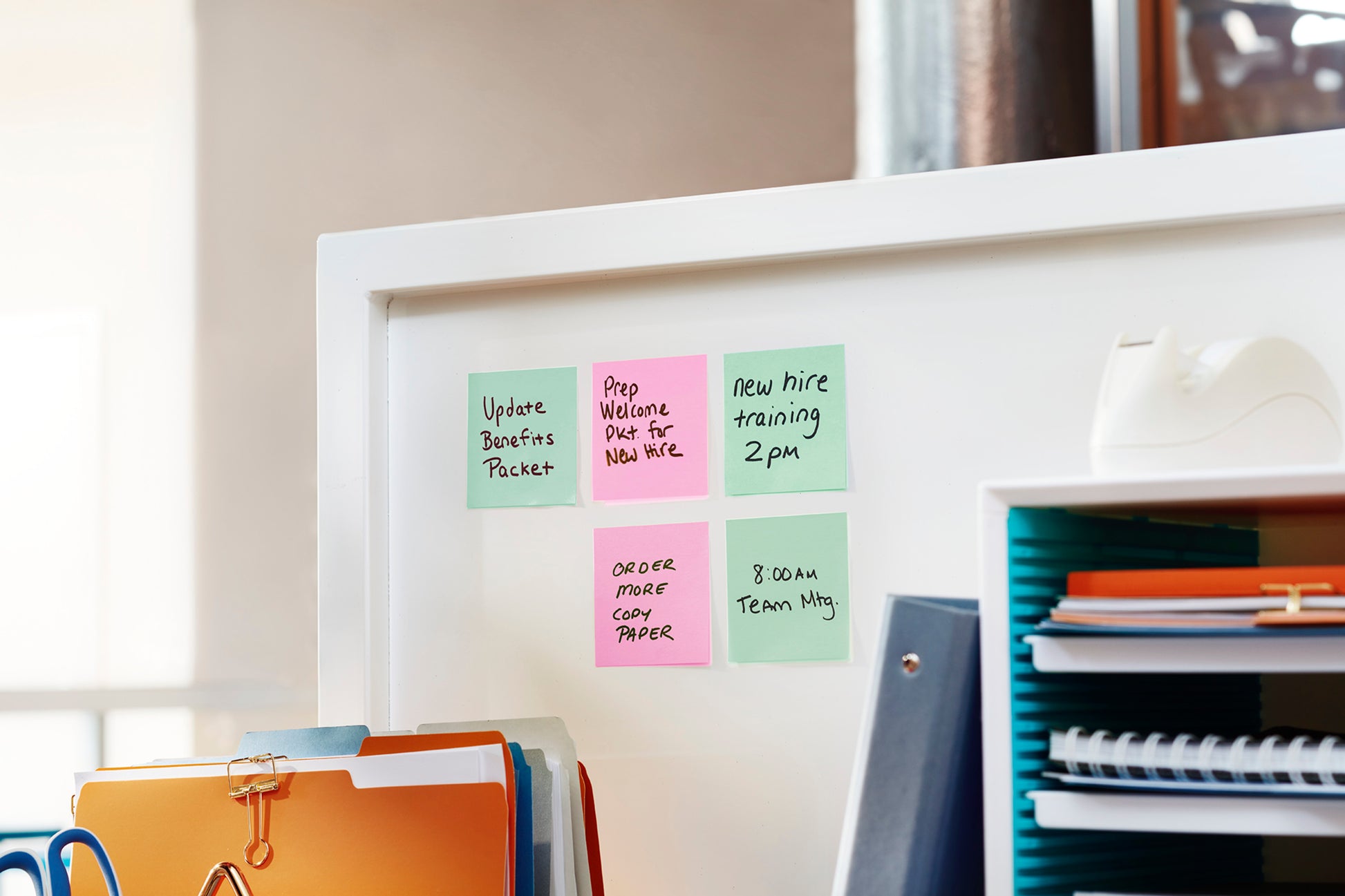Zwölf Blöcke der 3M Deutschland GmbH Post-it® Notes, Beachside Collection (38 mm x 51 mm), sind an einem weißen Schrank in einem modernen Arbeitsbereich angebracht, in dessen Nähe Ordner und Büromaterialien übersichtlich angeordnet sind.