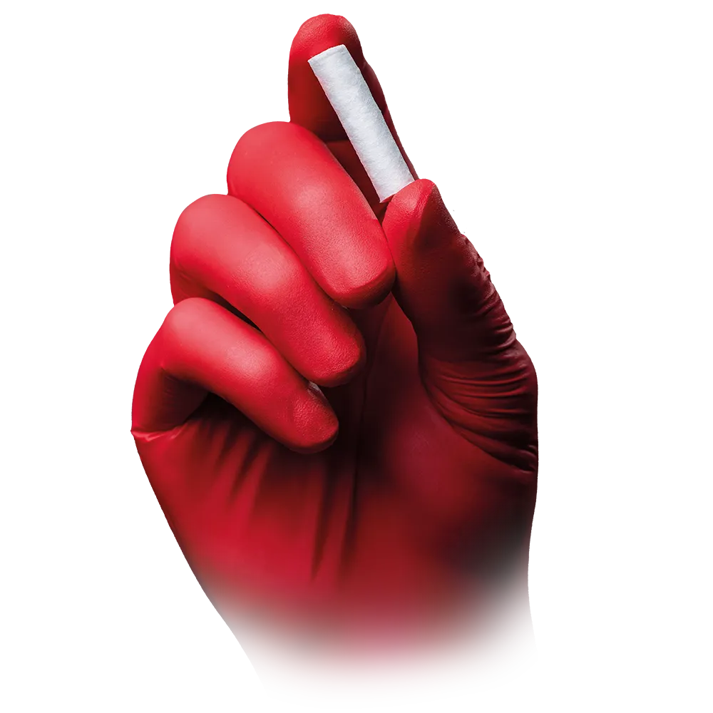 Eine mit einem roten Handschuh bekleidete Hand greift vor einem schlichten weißen Hintergrund vorsichtig ein kleines gerolltes Stück AMPri MED-COMFORT Dental Zahnwatterollen der AMPri Handelsgesellschaft mbH aus chlorfrei gebleichter Baumwolle zwischen Daumen und Zeigefinger.
