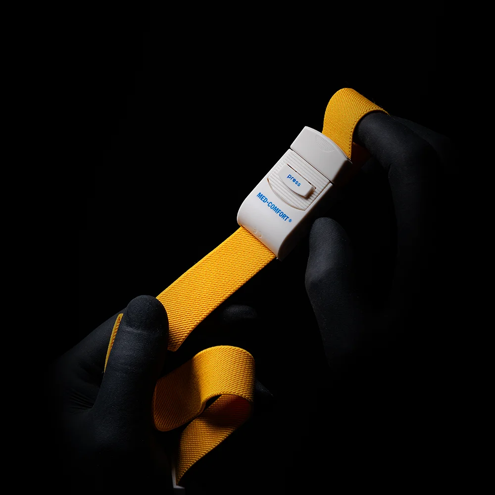Eine Person mit schwarzen Handschuhen hält einen AMPri MED-COMFORT Venenstauer, eine gelbe Aderpresse mit weißer Schnalle, zentriert vor schwarzem Hintergrund.