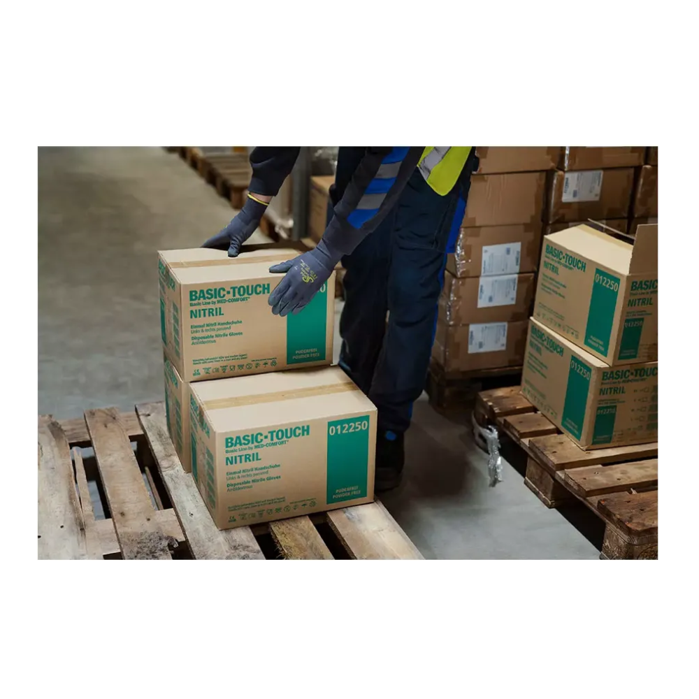 Eine Person, die AMPri SolidSafety Tough Micro Touch ultraleichte Montagehandschuhe trägt, stapelt sorgfältig Kartons mit der Aufschrift „Basic-Touch Nitrile“ auf einer Holzpalette in einem Lagerhaus. Mehrere andere Kartons sind sichtbar, und die Person scheint den Bestand mit Präzision und Sorgfalt zu ordnen.