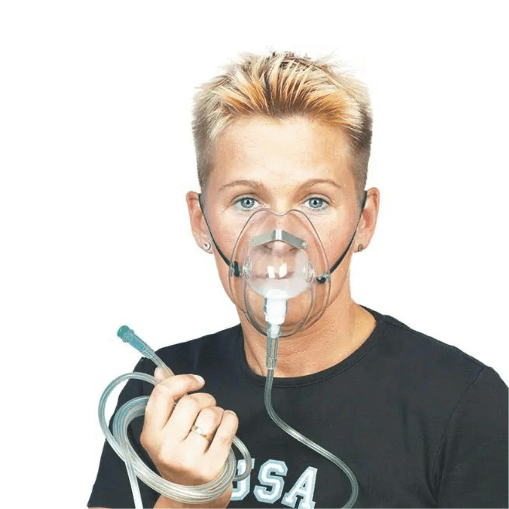 Eine Person mit kurzen blonden Haaren trägt die DCT Sauerstoffmaske für Erwachsene der Servoprax GmbH und hält den daran befestigten O2-Schlauch in der Hand. Sie trägt ein schwarzes Shirt mit weißer Schrift auf schlichtem weißen Hintergrund.