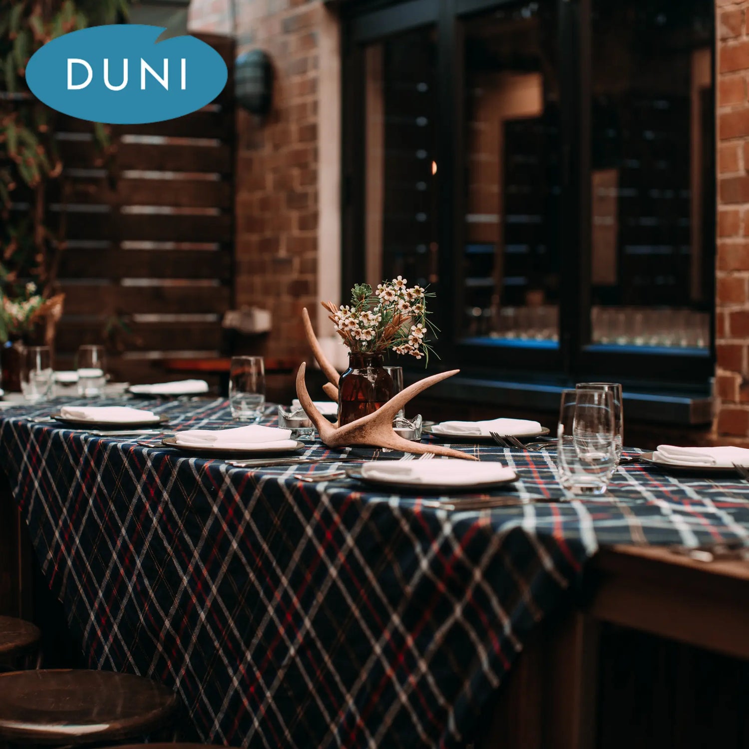 Ein gedeckter Tisch mit einem karierten Tischtuch, einer Vase mit Blumen und Hirschgeweihen als Dekoration, umgeben von Gläsern und Tellern, begleitet vom Duni-Logo im oberen linken Bereich